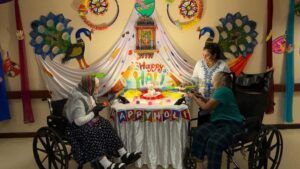 People in wheelchairs gather in front of a decorated table for a Holi celebration with colorful peacock wall art.