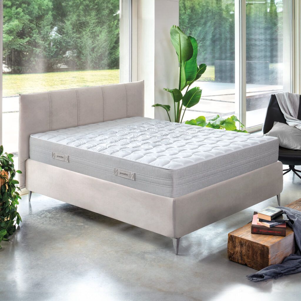 Letto beige con materasso bianco in una stanza ben illuminata, con una grande finestra e verde.