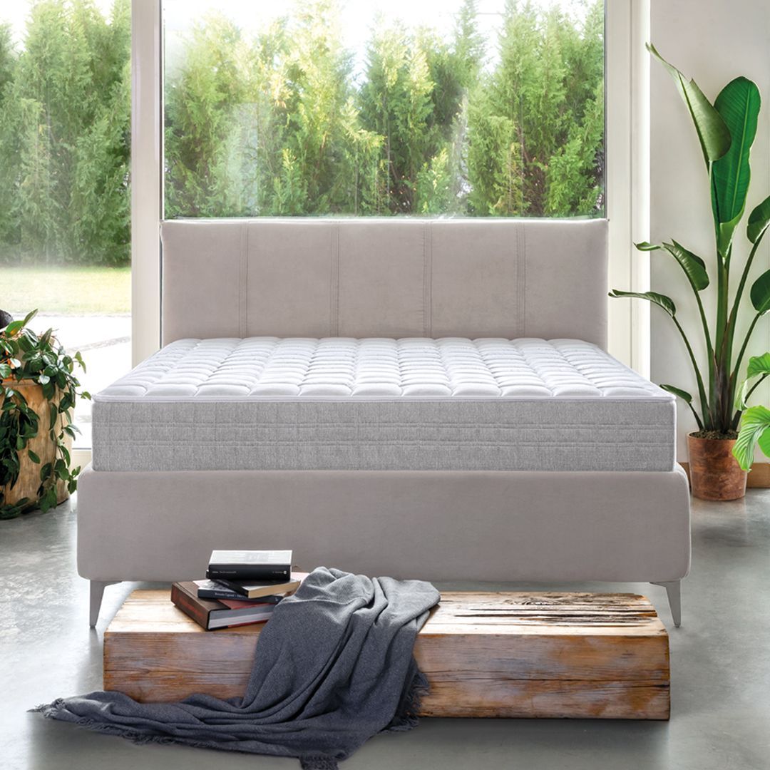 Letto beige in una stanza luminosa con una finestra che si affaccia su un lussureggiante paesaggio verde. Una panca di legno contiene libri e una coperta.