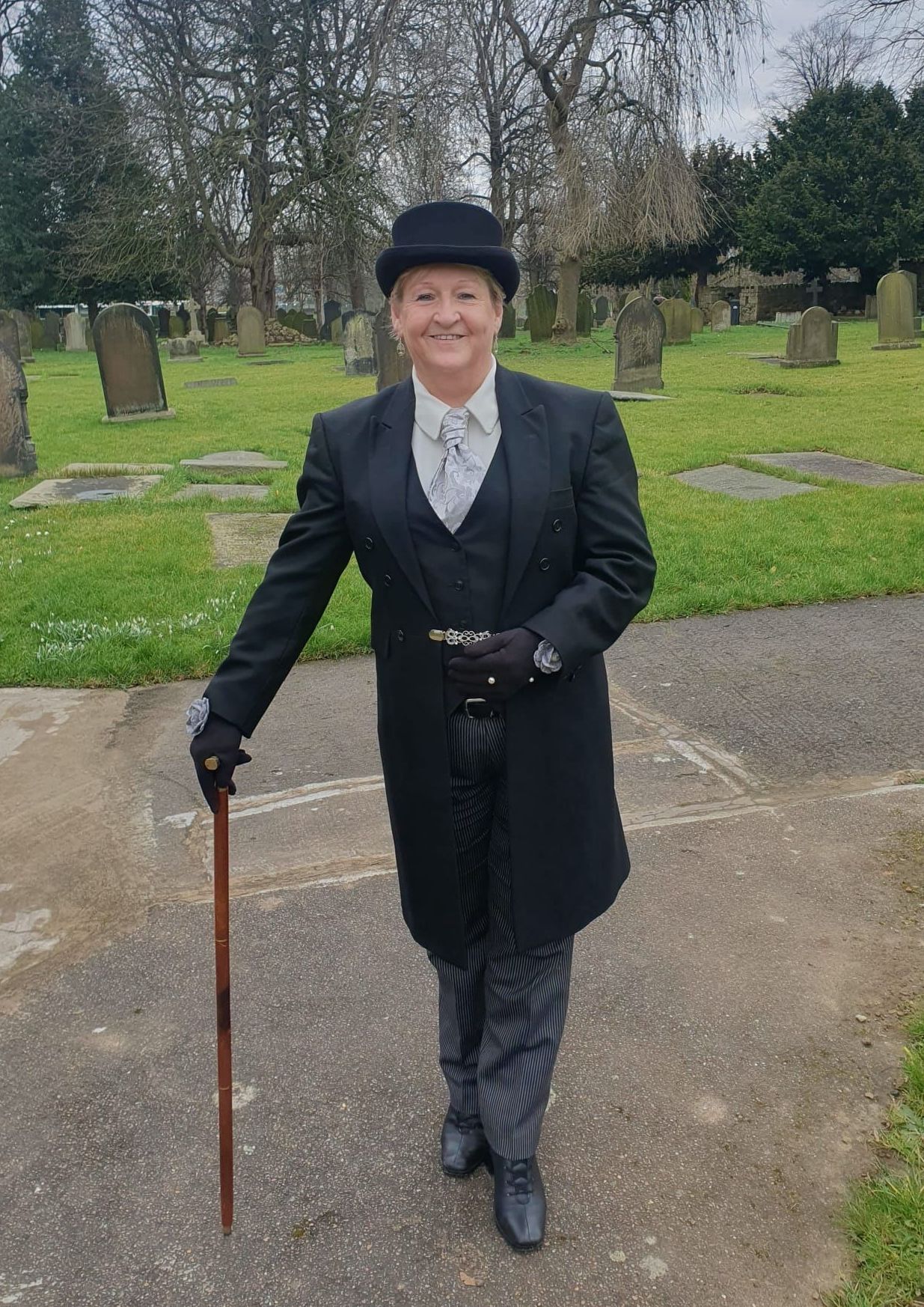 A man in a suit and top hat is holding a cane in a cemetery.