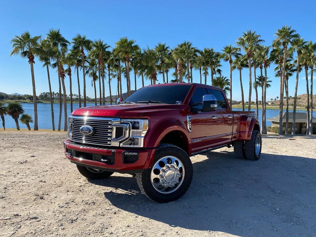A red truck is parked in a dirt lot next to a body of water.