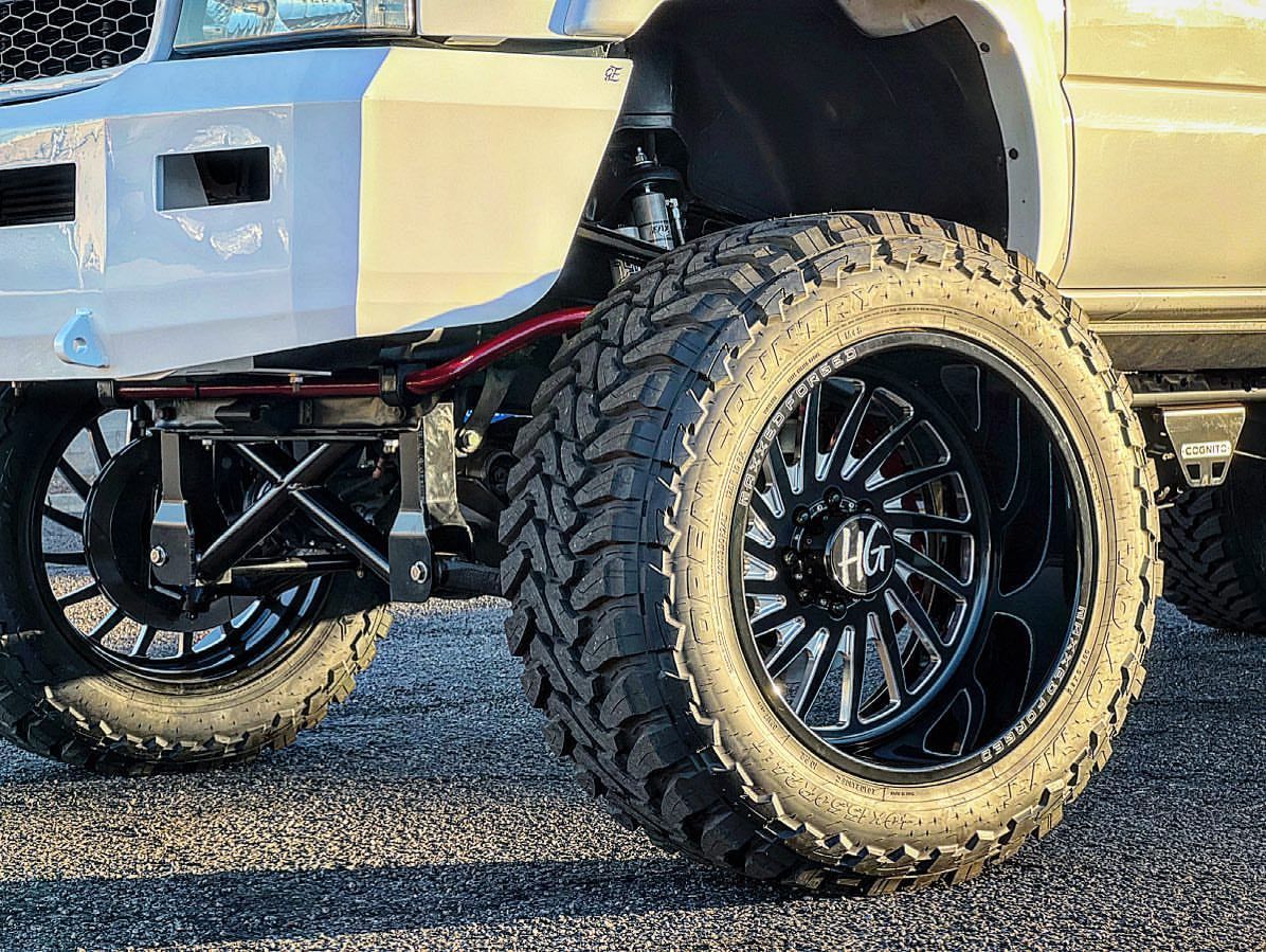 A white truck with black wheels and tires is parked on a gravel road.