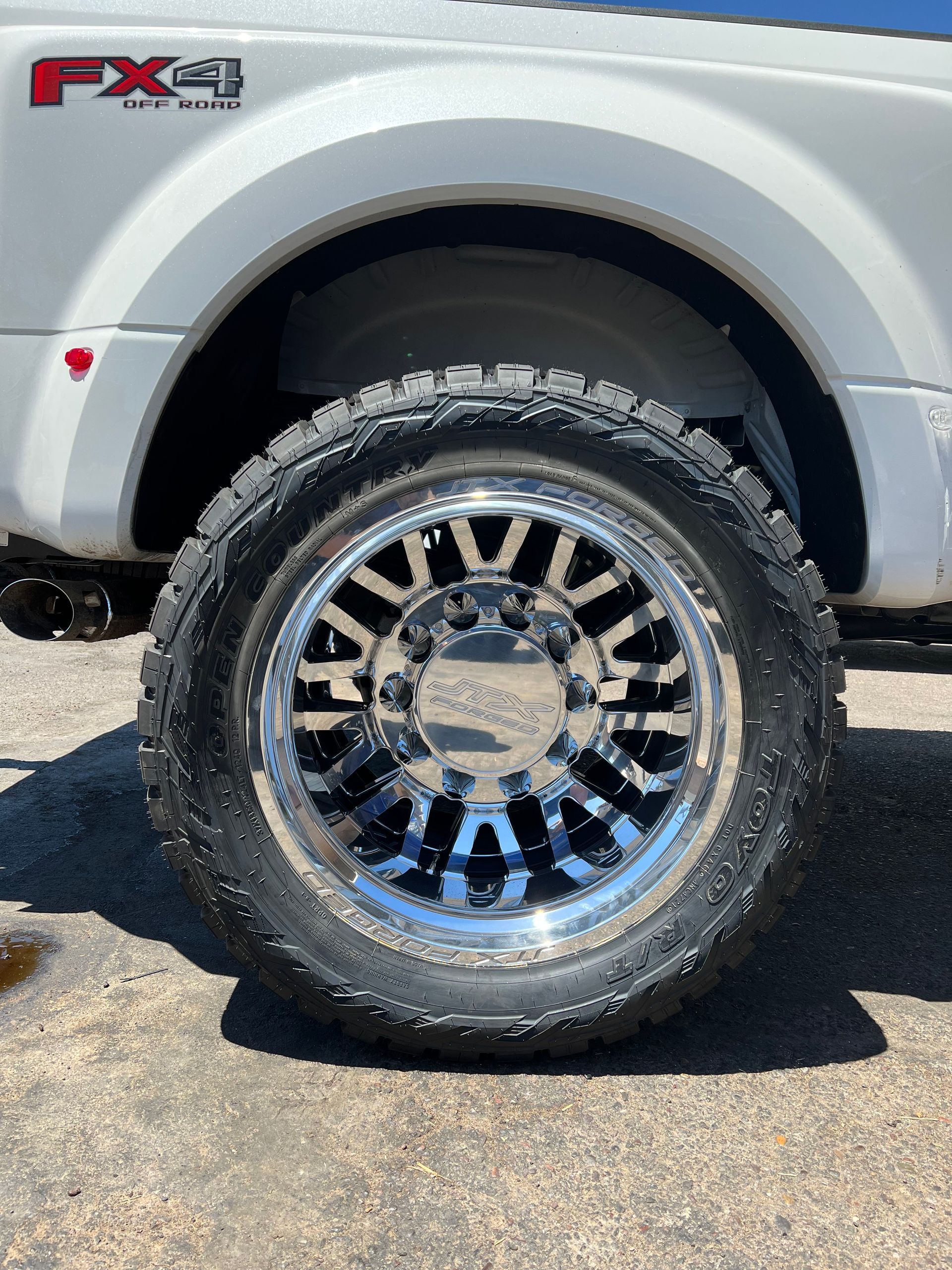 A white truck with a black tire and chrome wheels is parked on the side of the road.