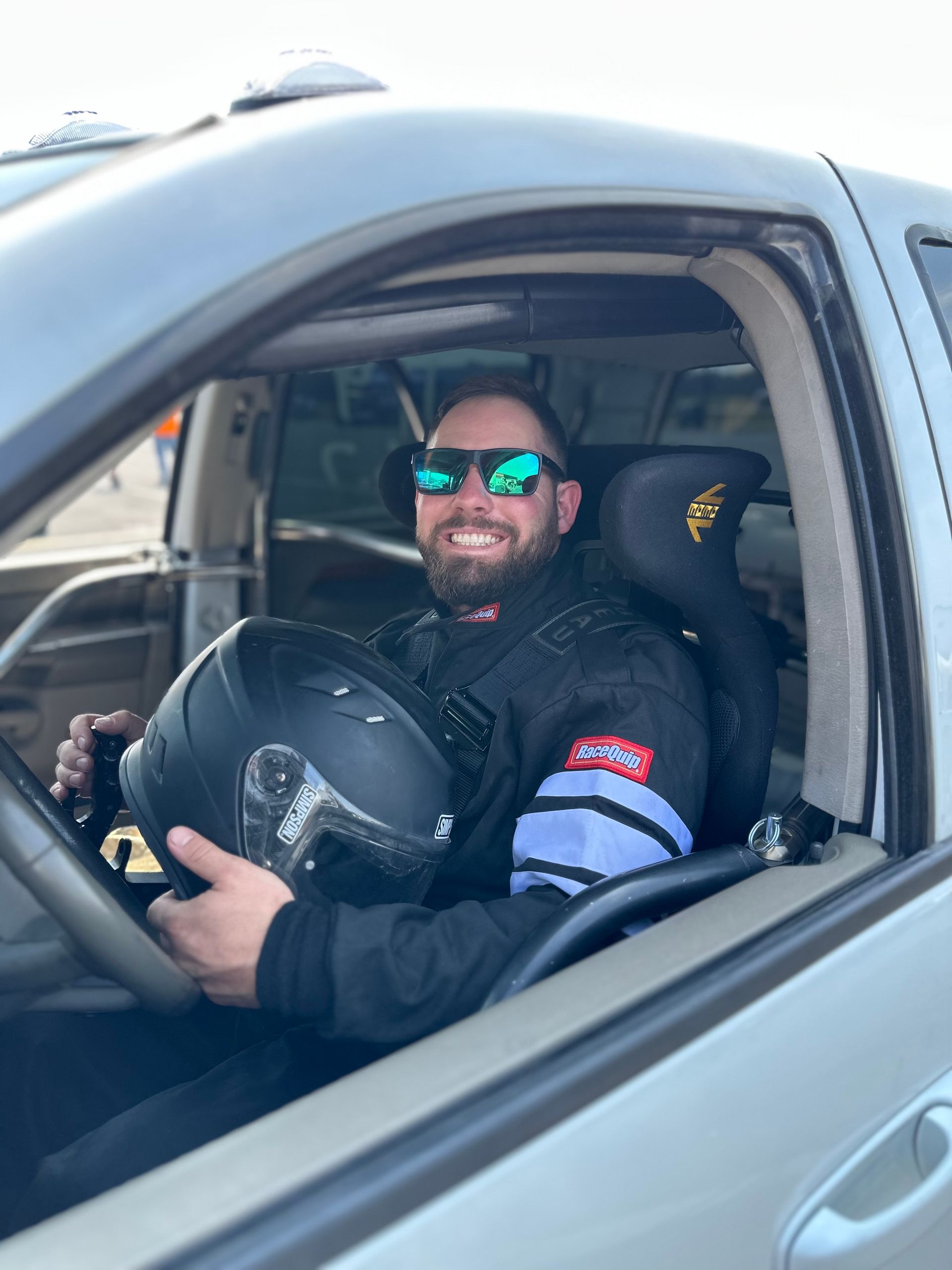 A man wearing sunglasses is sitting in a car holding a helmet.