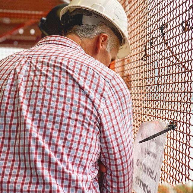 A man wearing a hard hat and a plaid shirt is looking at a clipboard.