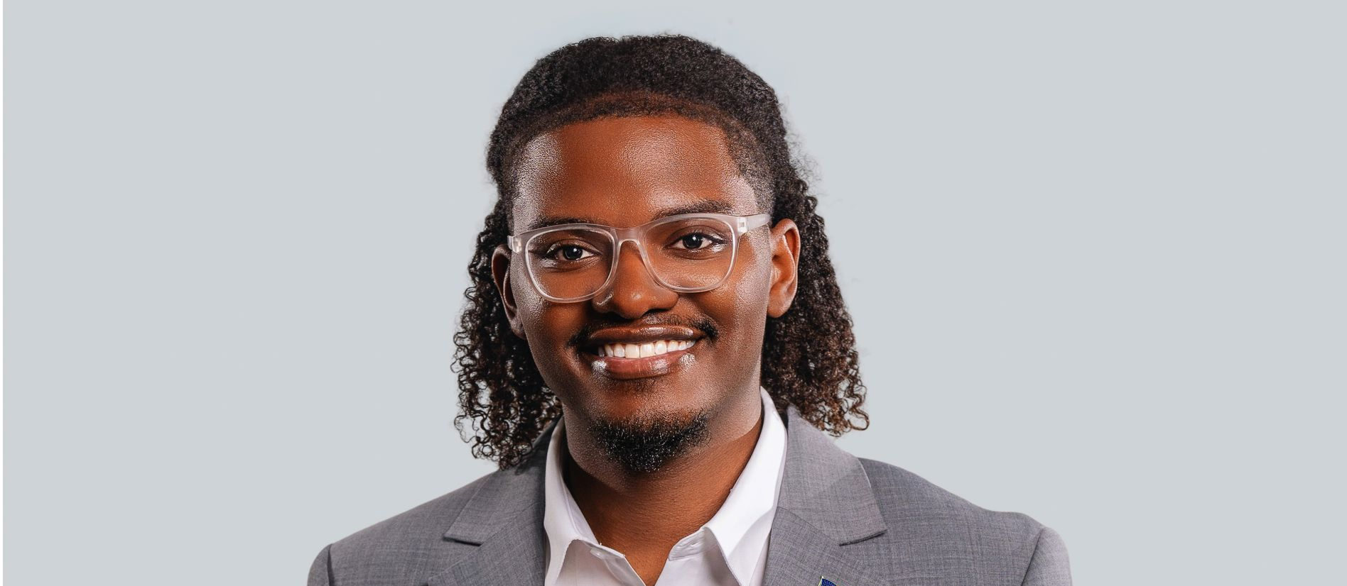 Man with glasses and long curly hair smiles in a suit jacket.