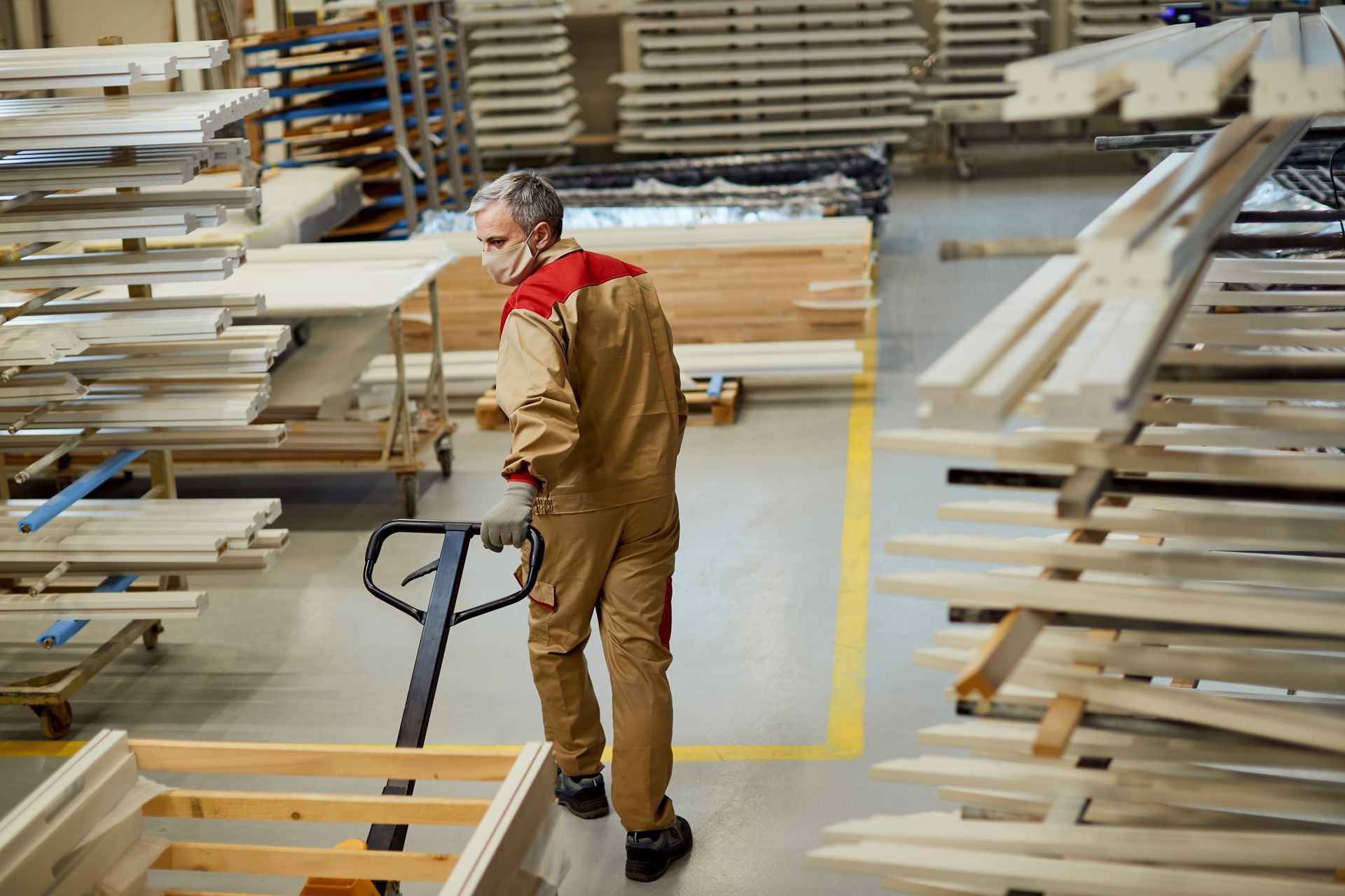 Worker in tan uniform steering a pallet jack through a lumber warehouse aisle