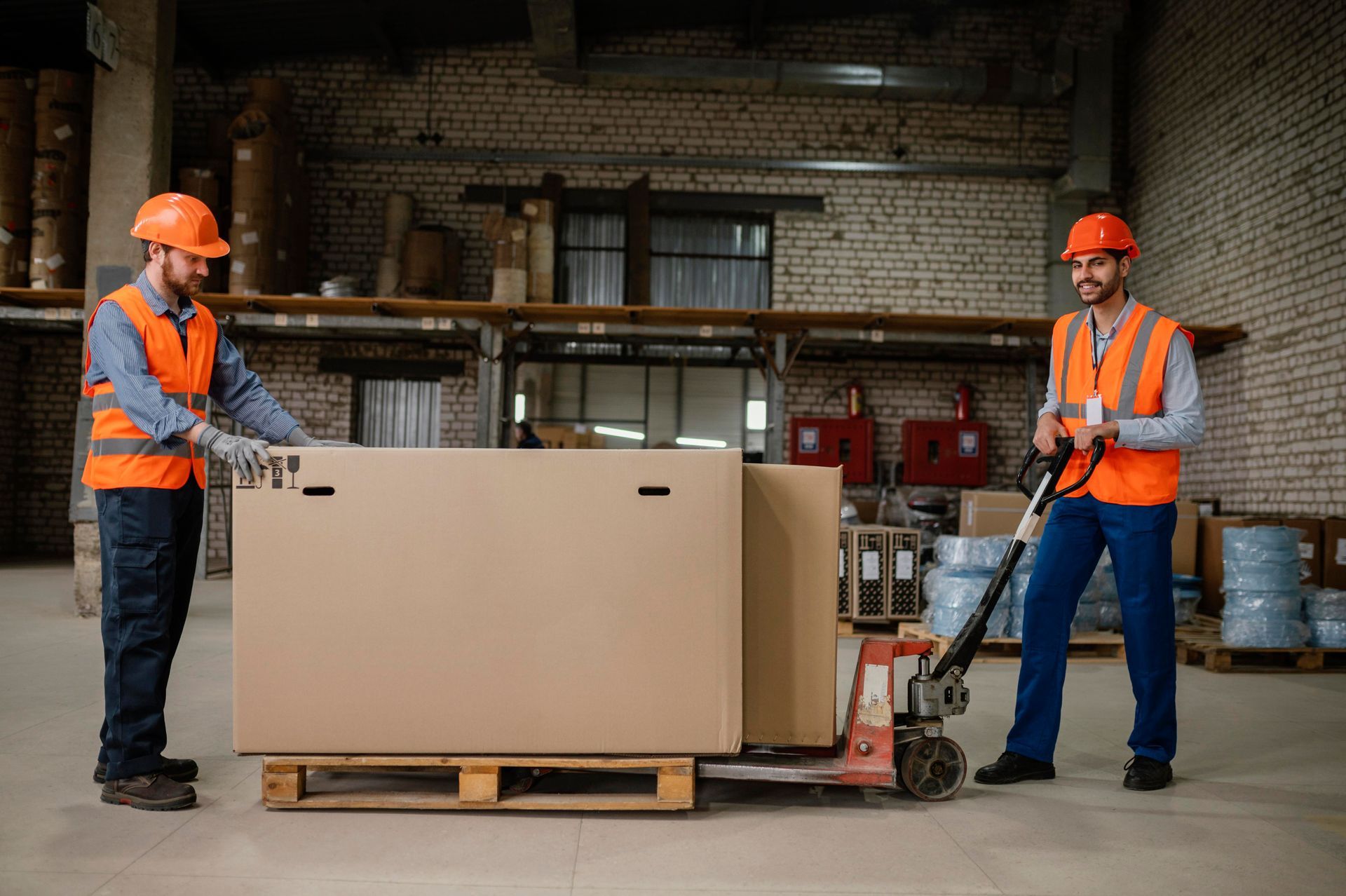 Two workers in orange safety gear move a large crate with a pallet jack in a warehouse.