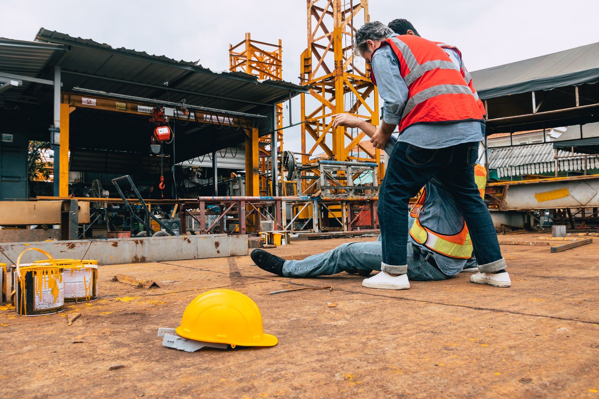 Construction worker on ground, another worker helping him. Hard hat in foreground, crane in background.
