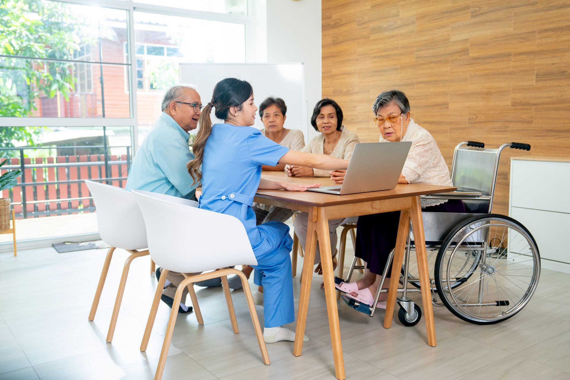 Nurse shows a laptop to a group of seated people at a table, one in a wheelchair, in a bright room.