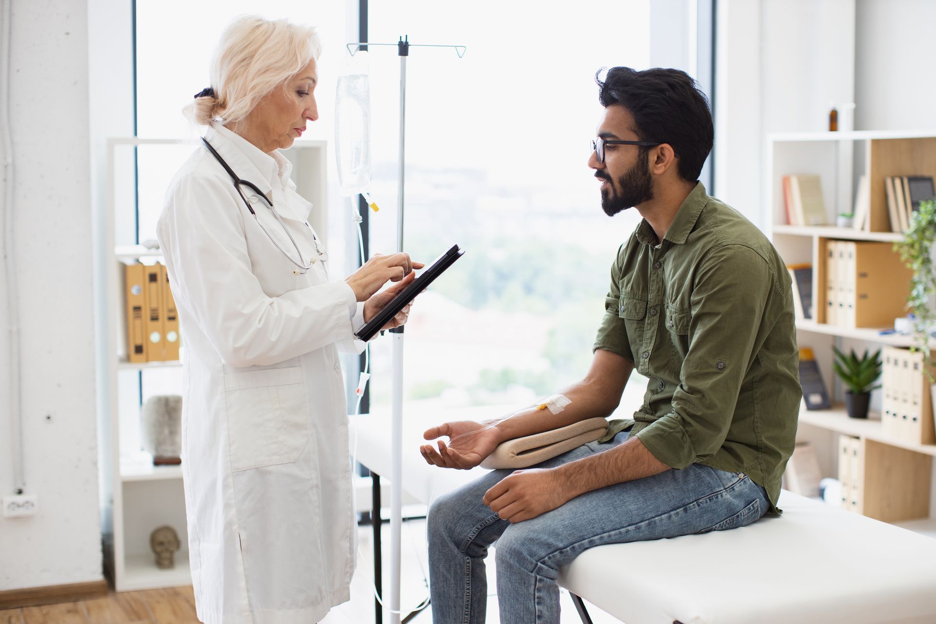 Doctor in white coat consults with a patient sitting on an exam table; IV in patient's arm, bright room.
