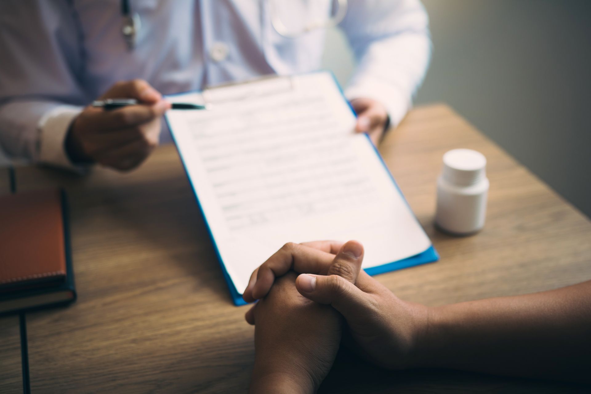 Doctor pointing at paperwork, patient's hands clasped on a table. White pill bottle in the background.