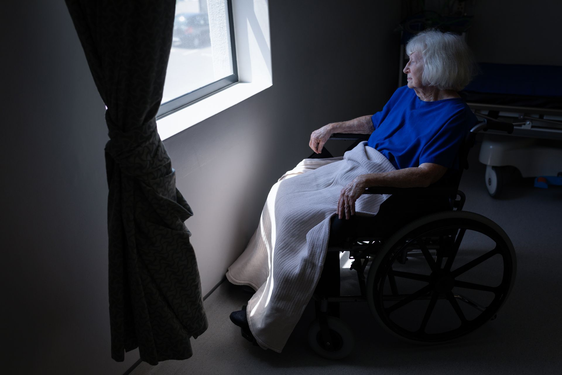 Woman in wheelchair looking out a window, blanket on lap, in a room with a hospital bed.