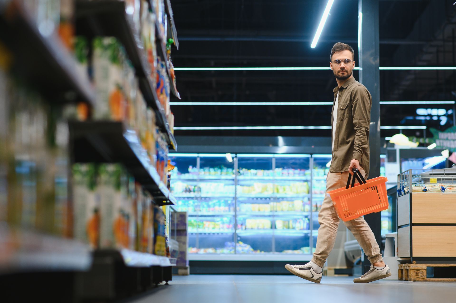 Man in store aisle holding orange basket, looking back. Refrigerated section in background.