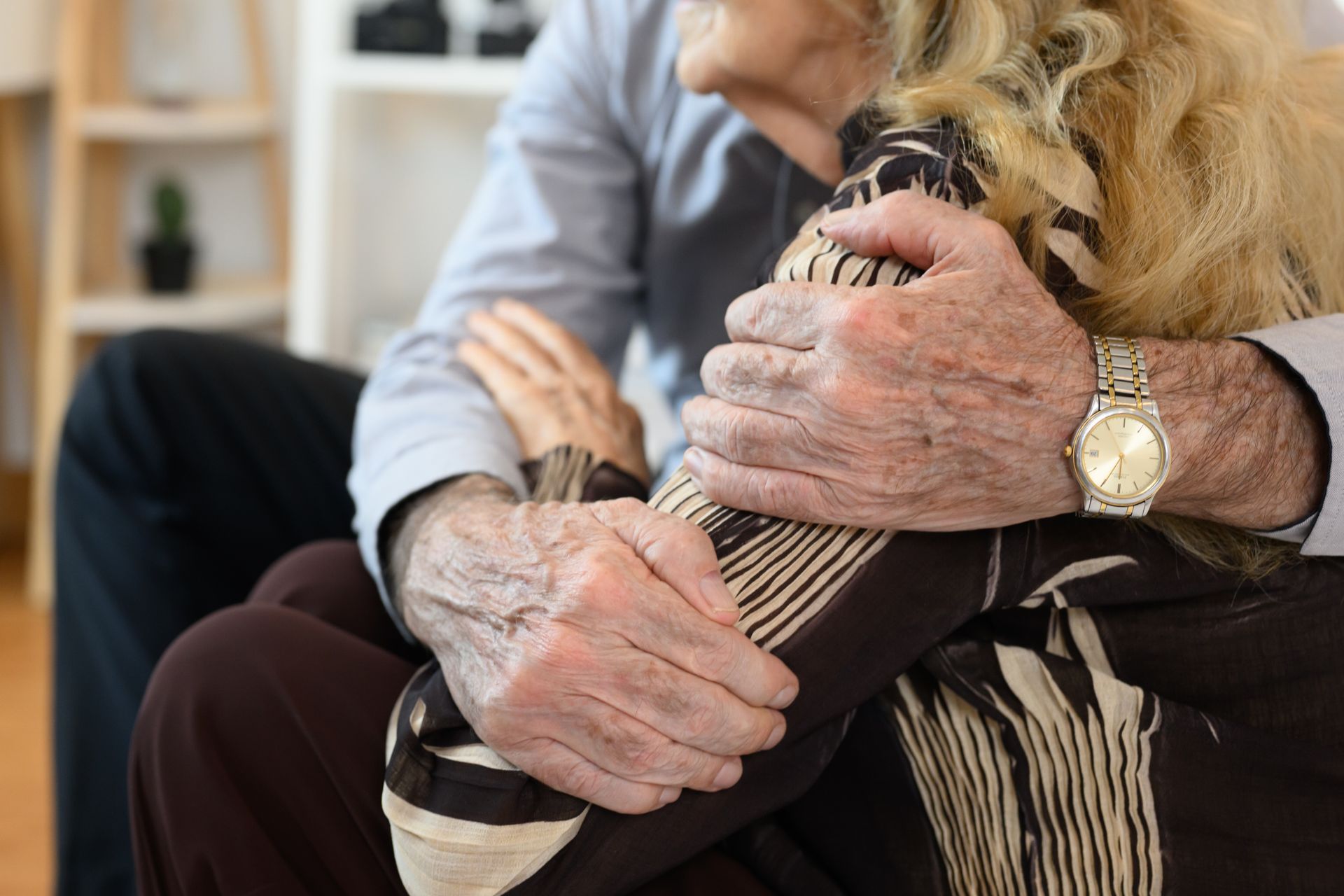Older person's hand with watch clasps arm of another person, brown patterned sleeve, in a seated position.