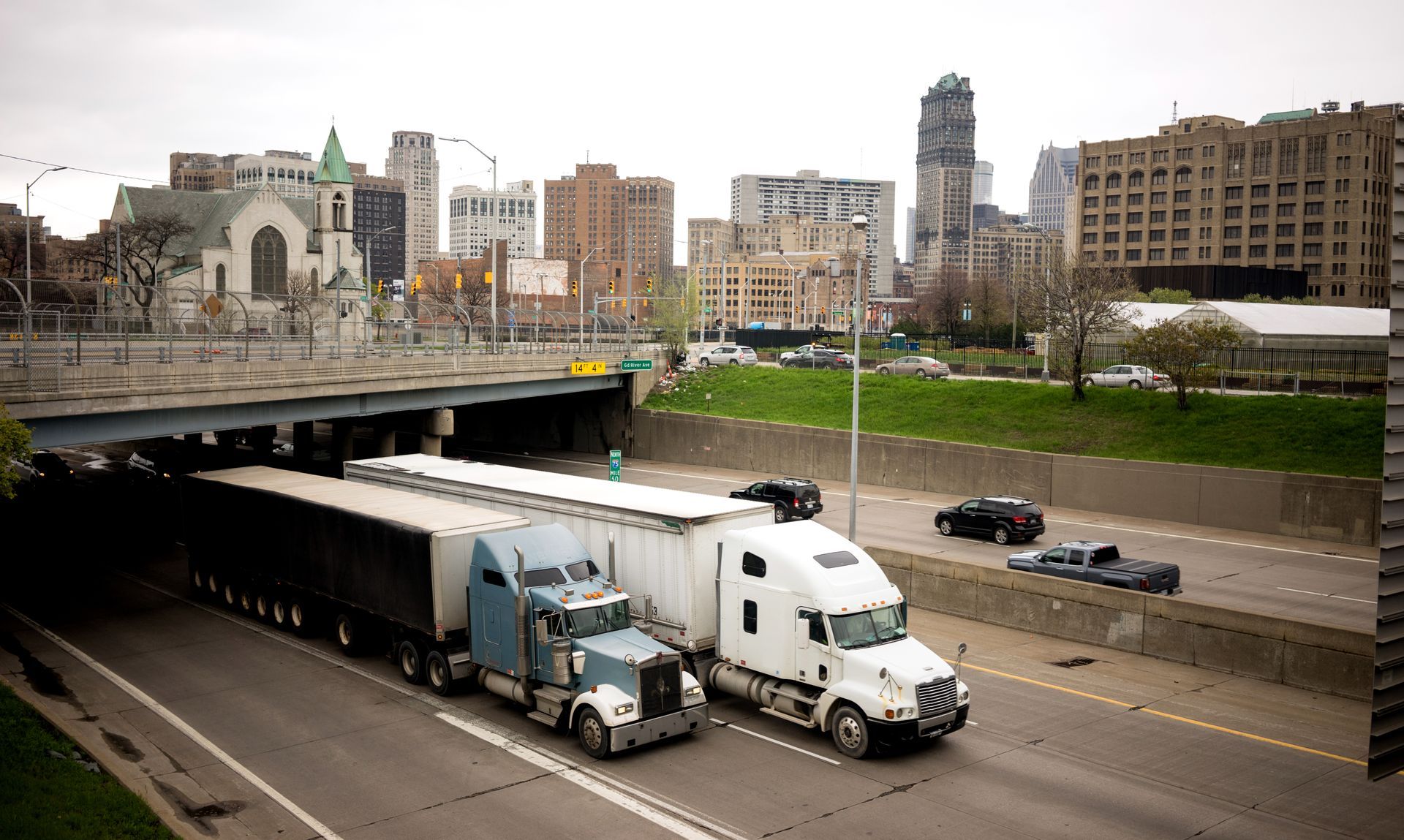 Trucks driving under an overpass in a city, buildings in the background. Overcast sky.