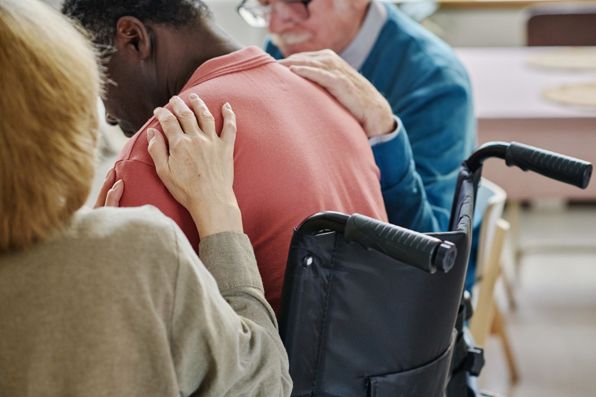 People comforting a person in a wheelchair, hands on his back; he is slumped over, appearing distressed.