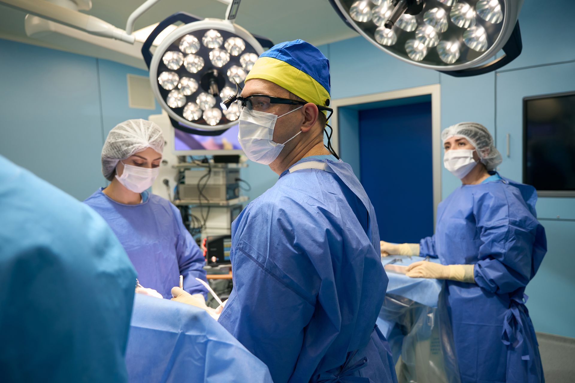 Medical team in blue scrubs, surgical masks, and caps operating under bright lights.