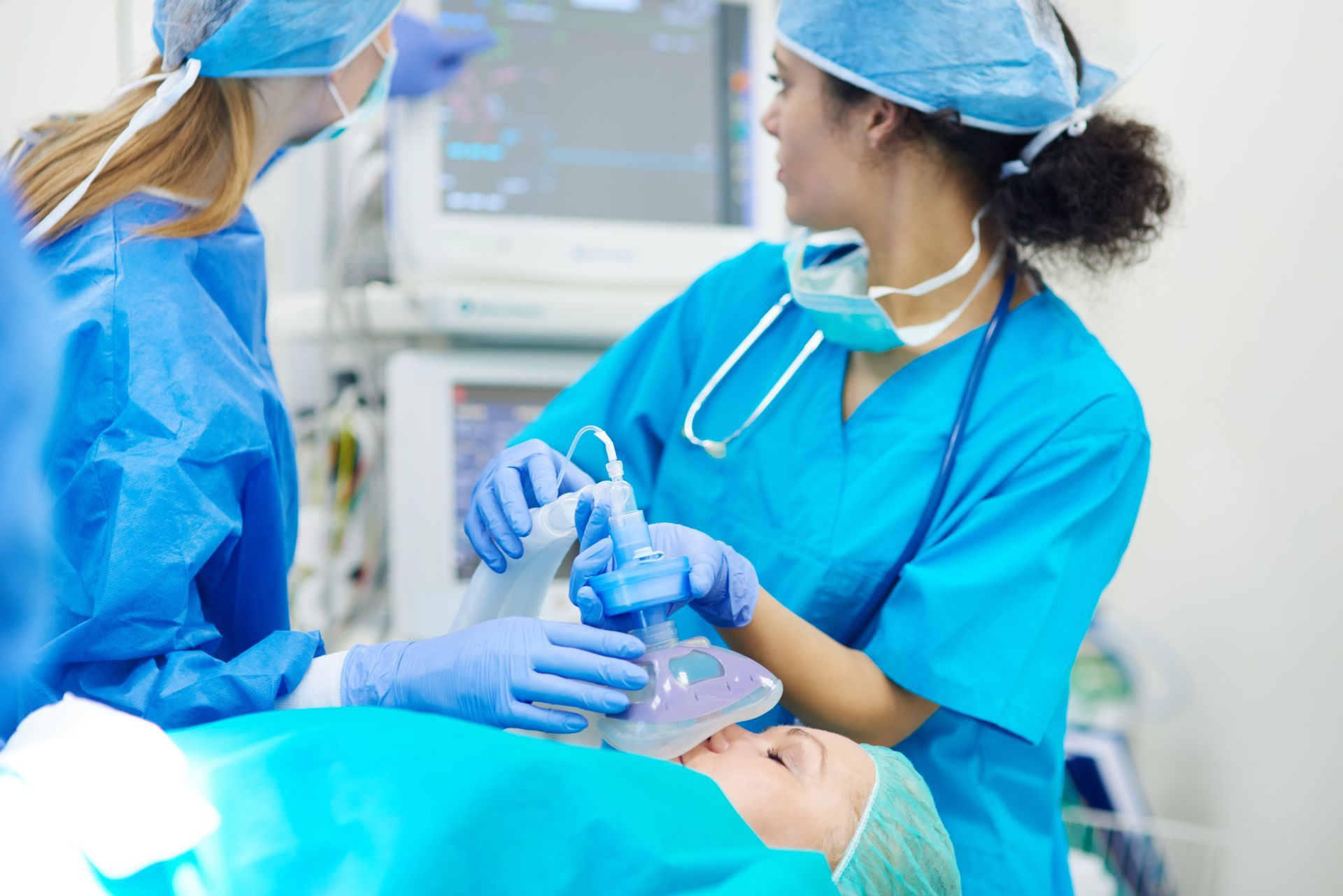 Medical personnel administer anesthesia to a patient in an operating room, monitors visible.