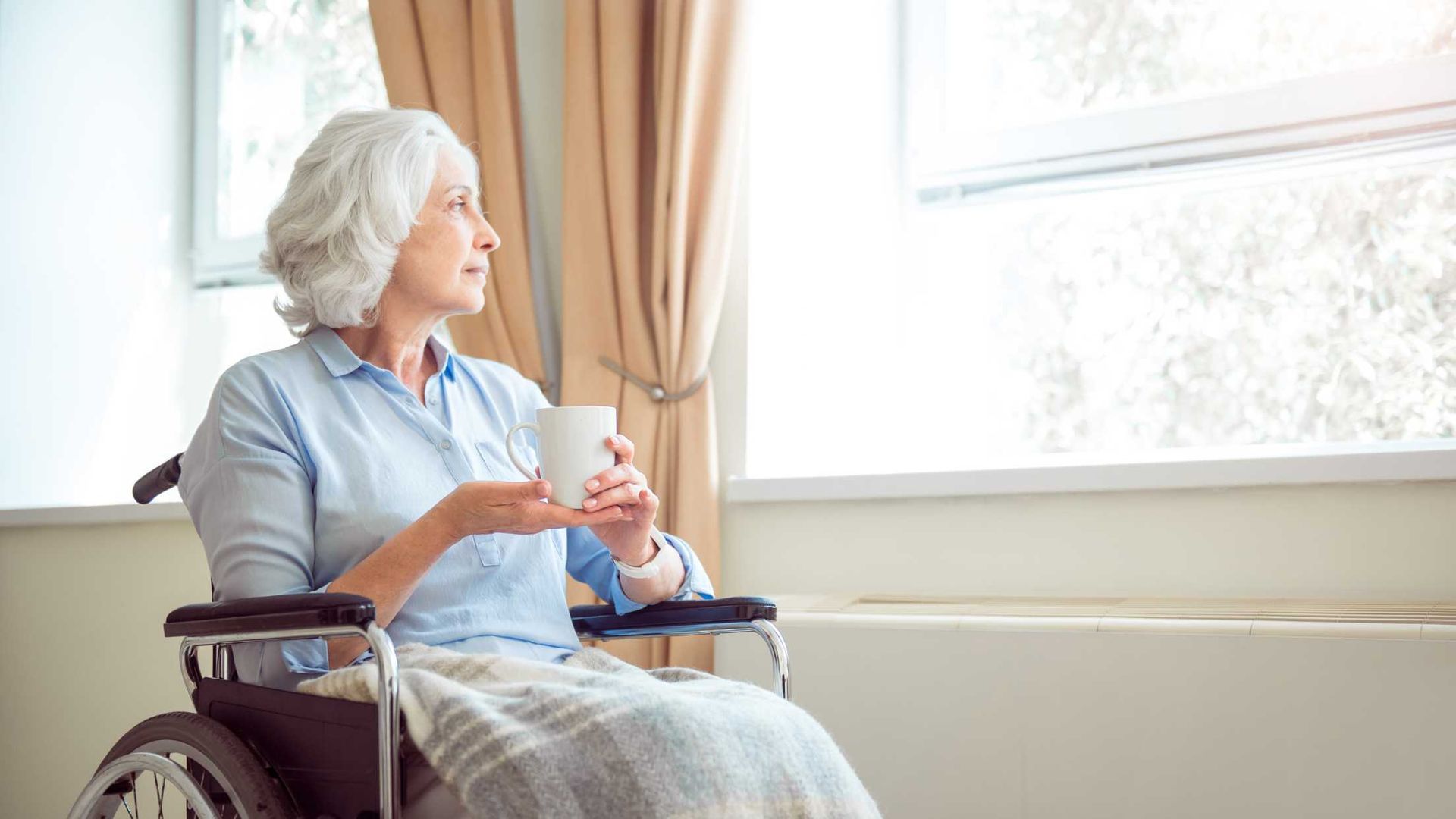 An elderly woman in a wheelchair is drinking coffee and looking out the window.