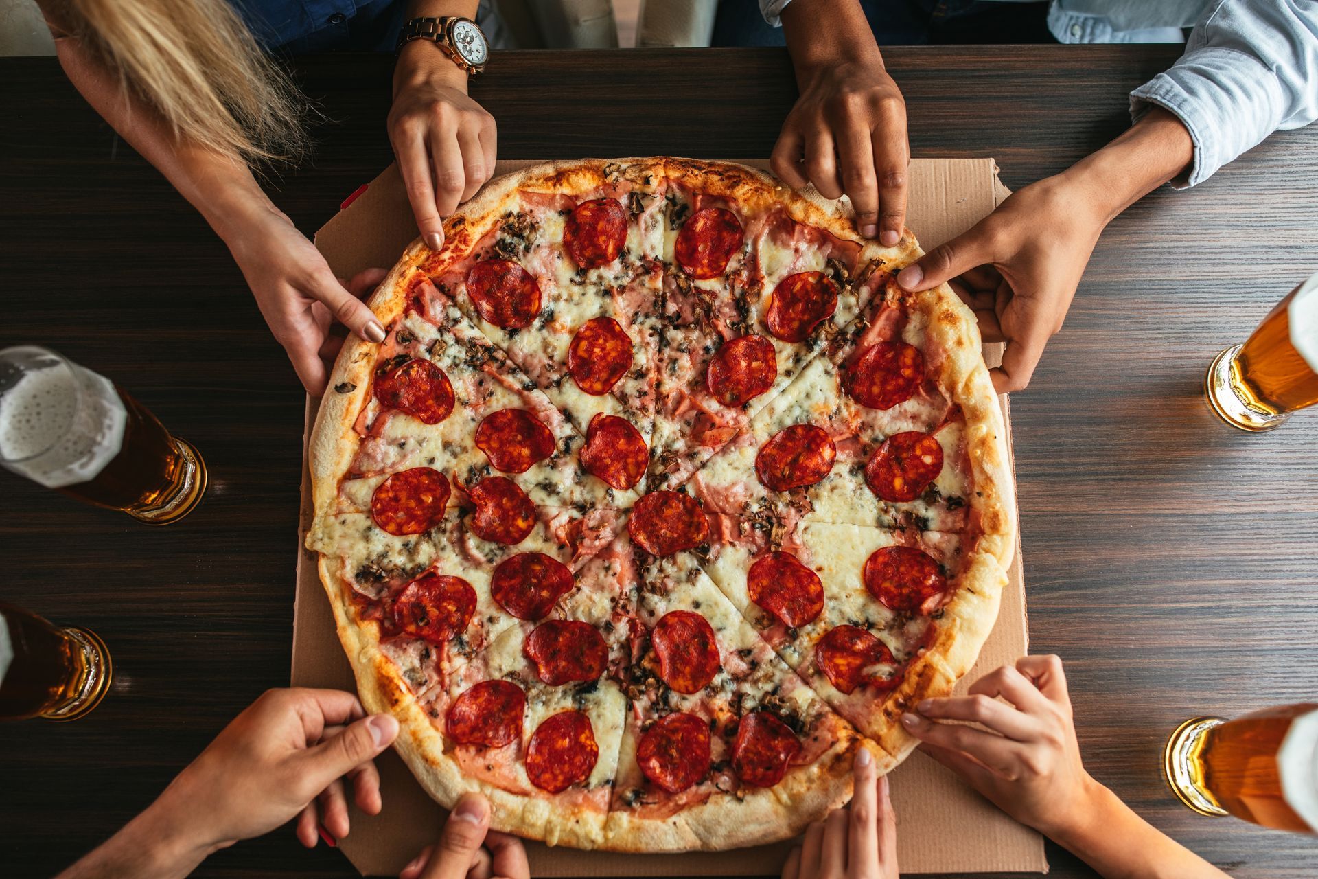 Hands reaching for a pepperoni pizza on a wooden table with drinks nearby