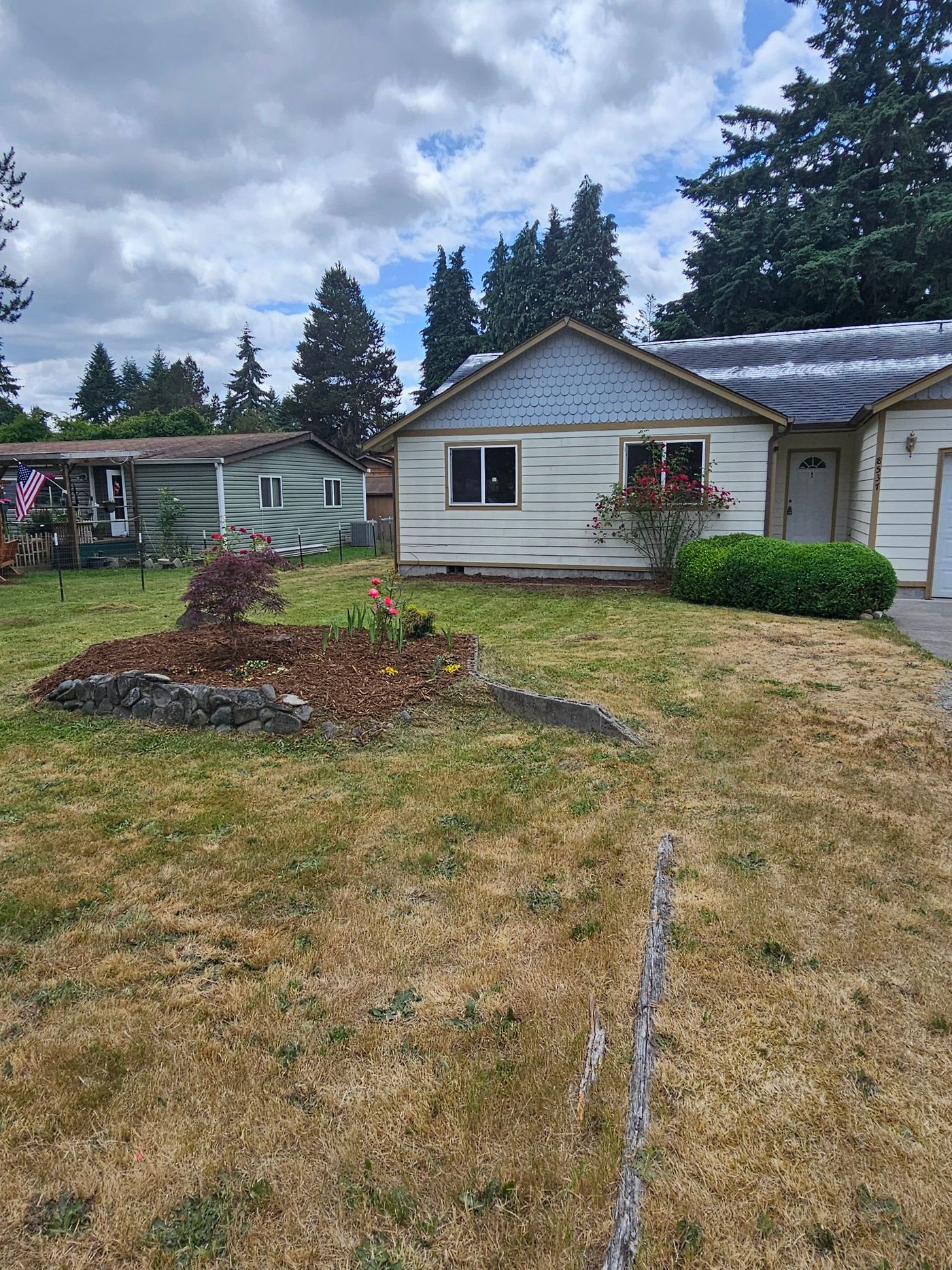 A weathered, single-story house with dry lawn, small flower bed, and partly cloudy sky.
