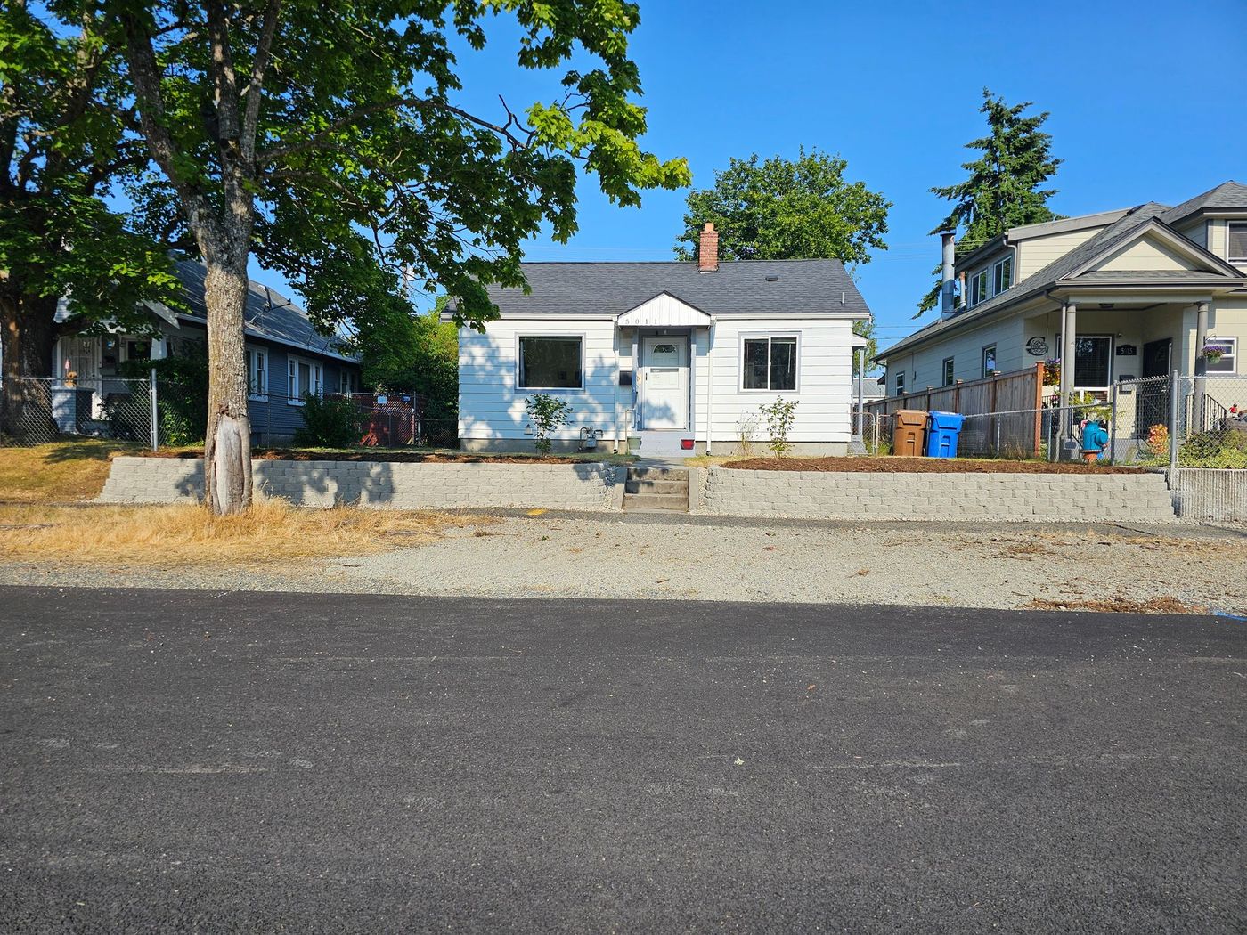 White house with grey retaining wall, trees, and asphalt road on a sunny day.