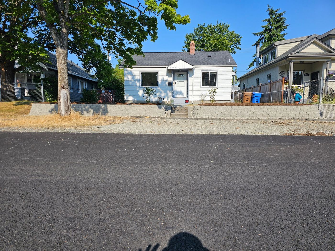 Small white house with a gray roof sits behind a concrete wall, next to a road with houses on either side.