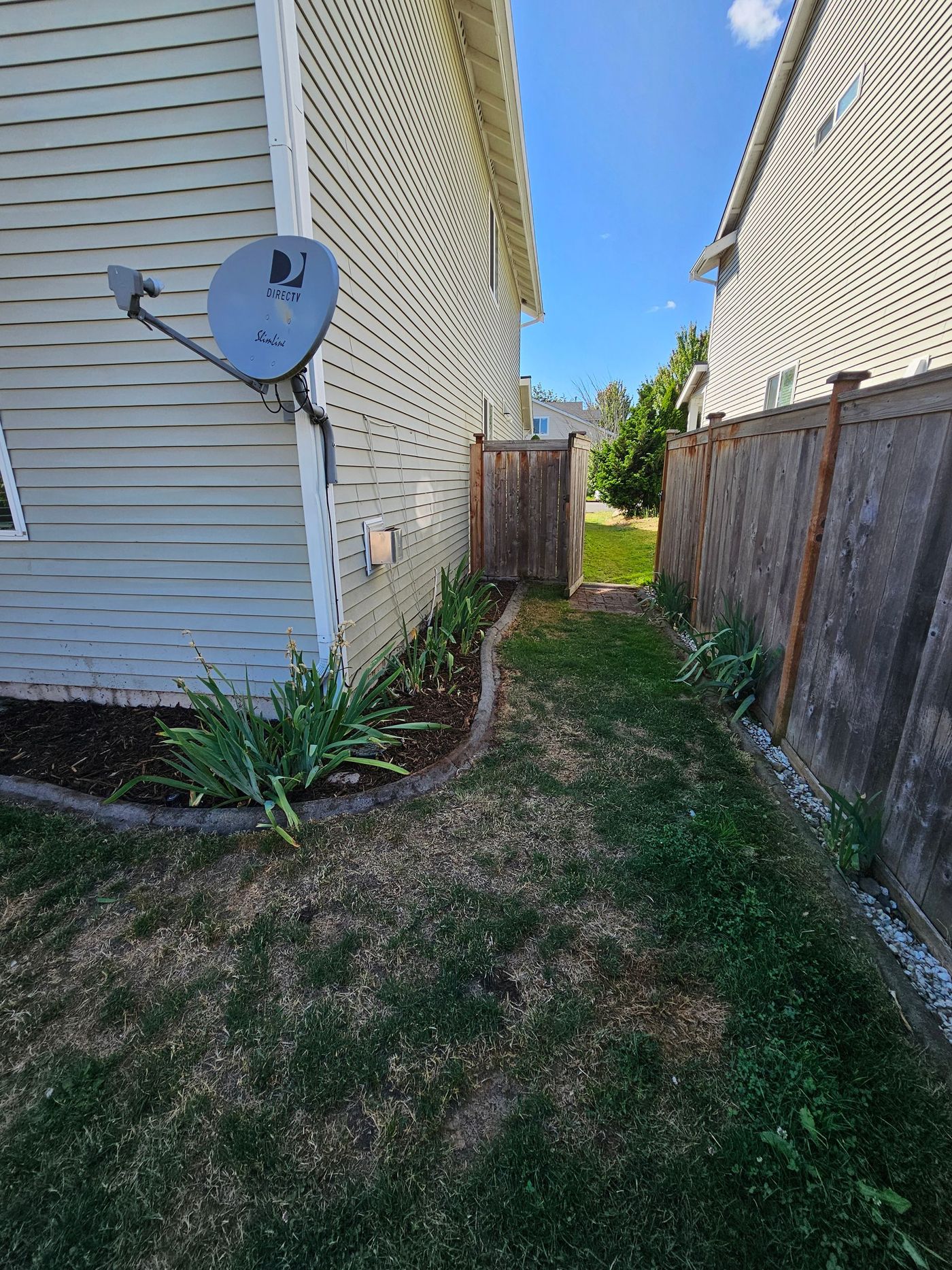Narrow grassy path between two houses; wooden fence and gate lead to a yard. Blue sky overhead.