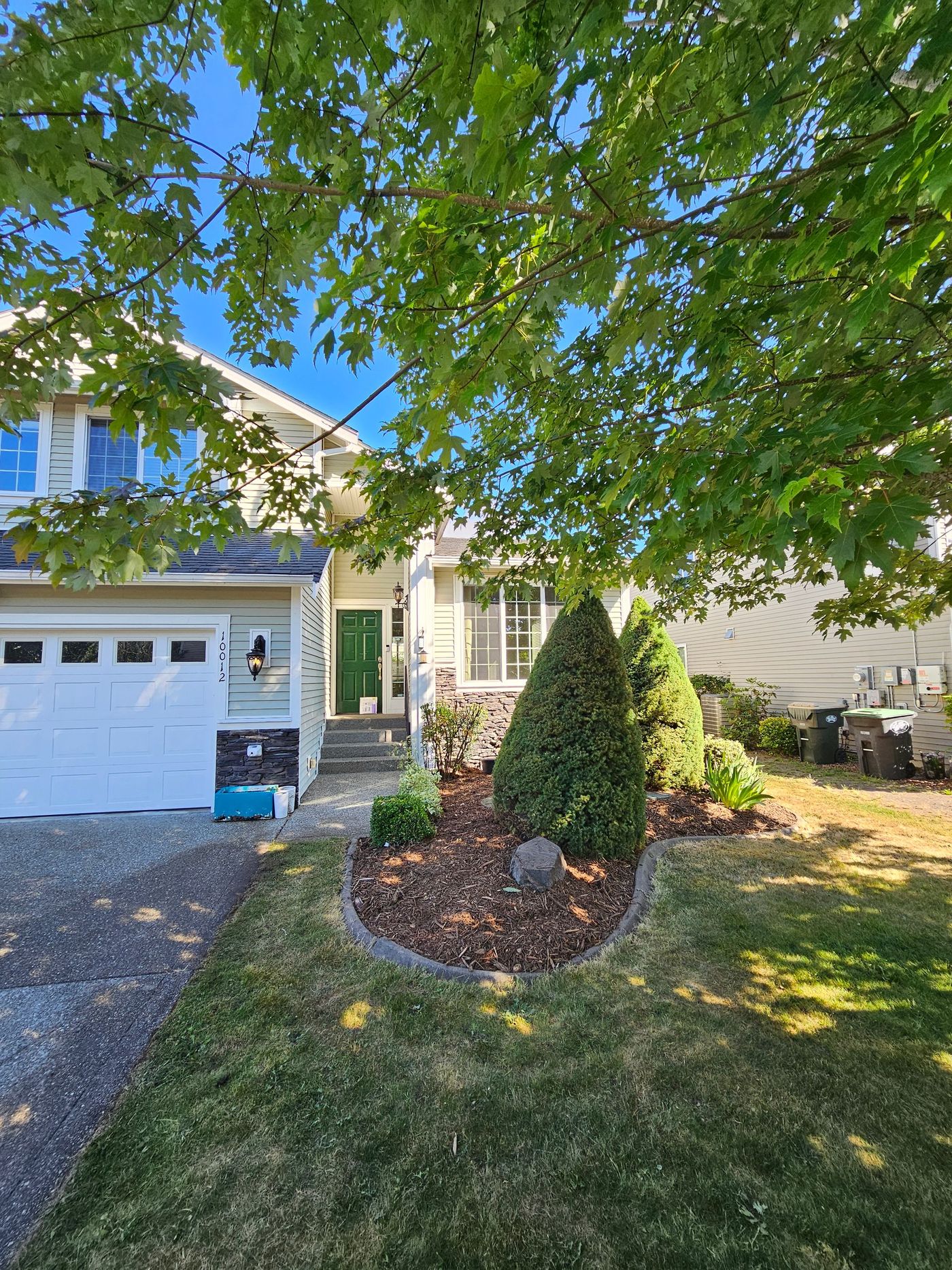 A house with a white garage, green door, and landscaped front yard, under a leafy tree.