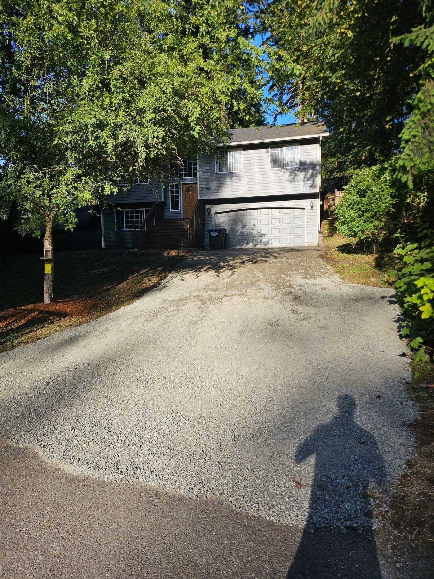 Gravel driveway leading to a two-story house with a garage, shadowed figure in foreground, trees on either side.