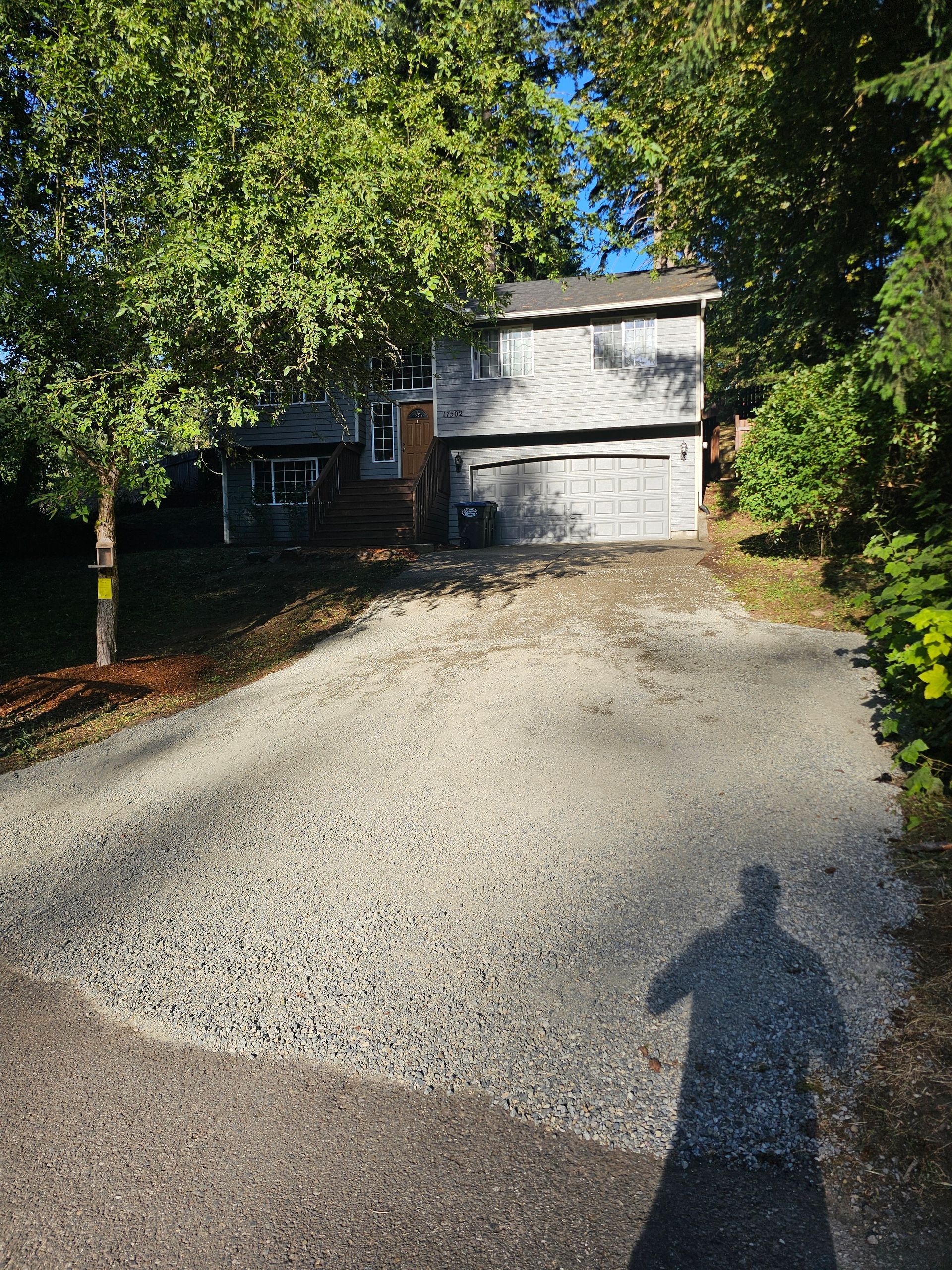 Gravel driveway leads to a two-story house with a garage. Shadow of a person in foreground.
