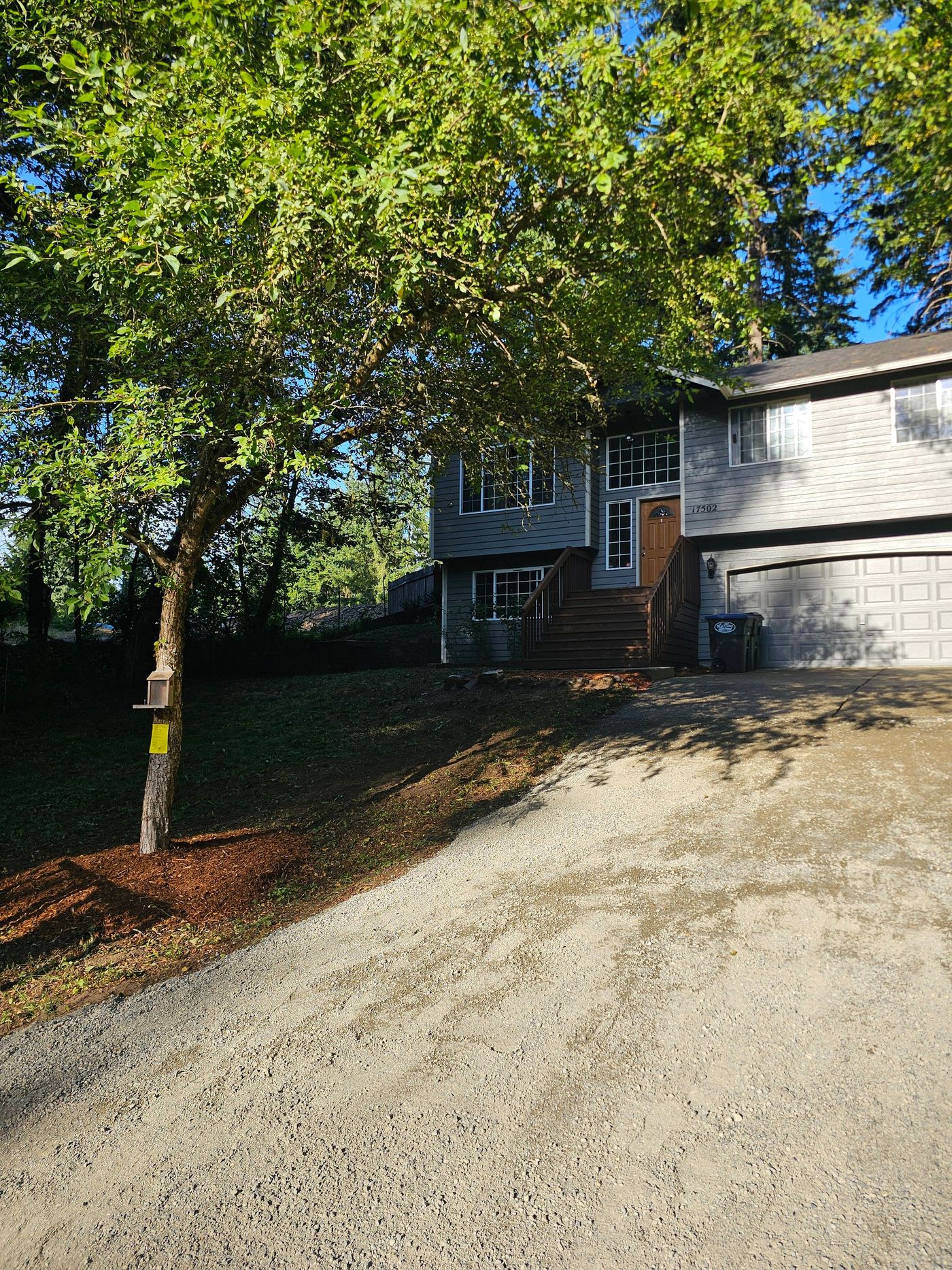 House with grey siding and dark roof, accessed by wooden stairs and gravel driveway, shaded by a large tree.