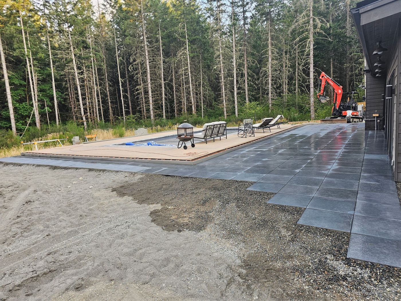 Patio construction: gray pavers and gravel, with pool area and excavator in wooded setting.