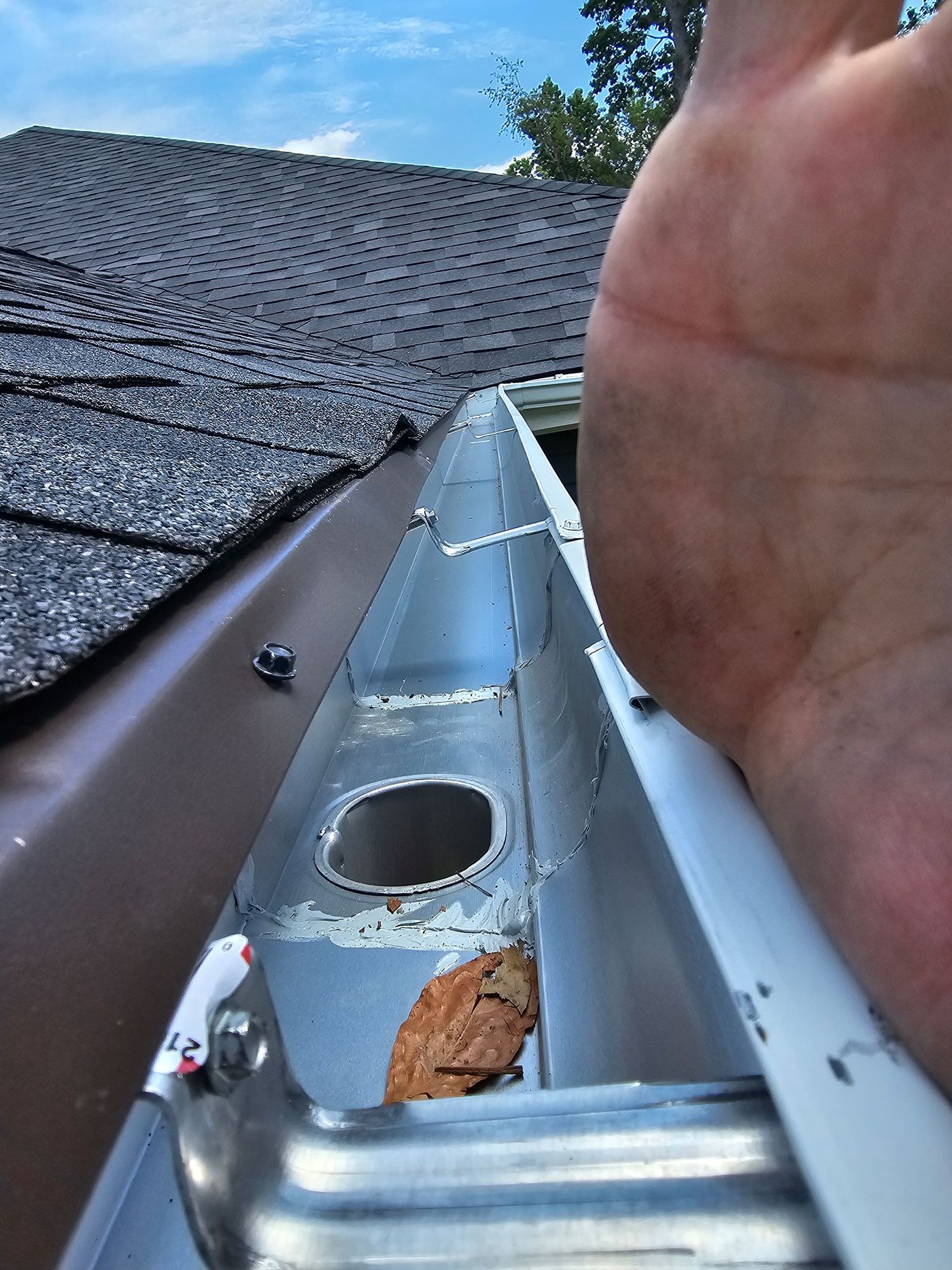Gutter with debris, brown roof, hand reaching in, blue sky.