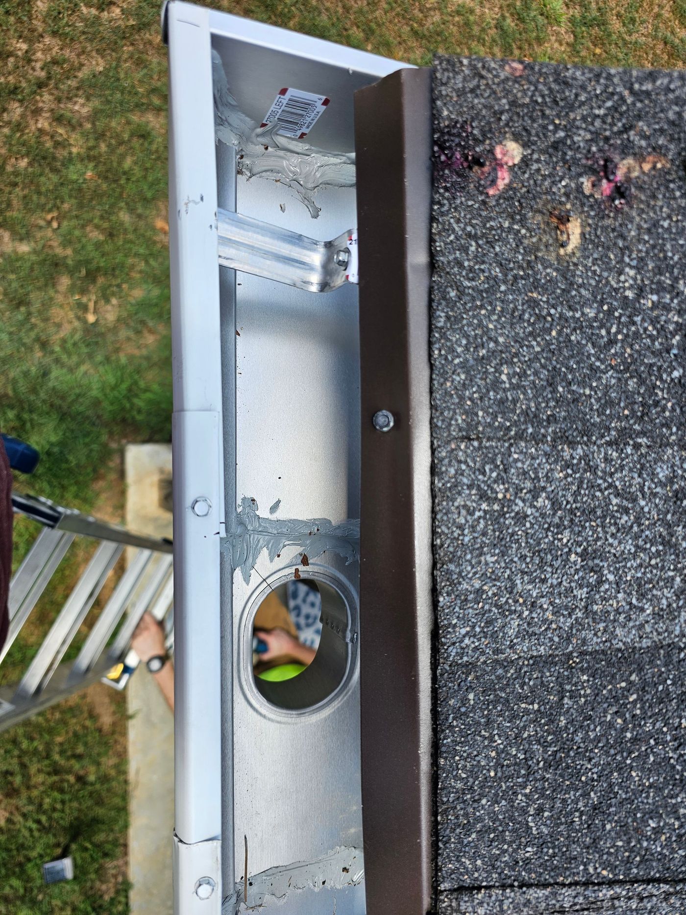 Section of a metal gutter with a drain hole next to a shingled roof.