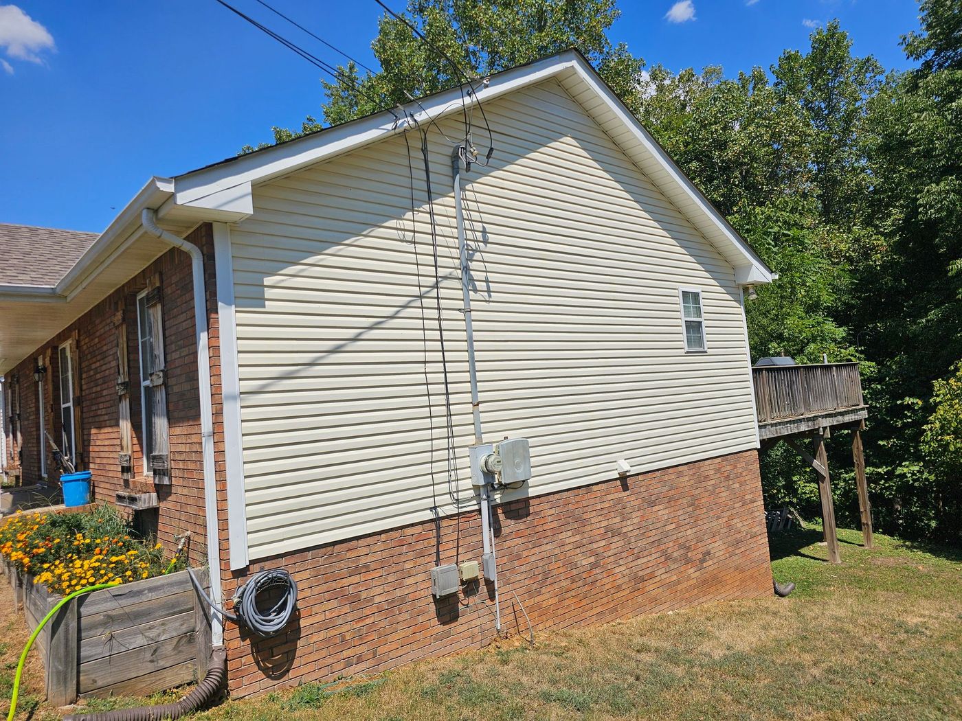 Side view of a house with tan siding, brick base, and a small wooden deck, set against a blue sky.