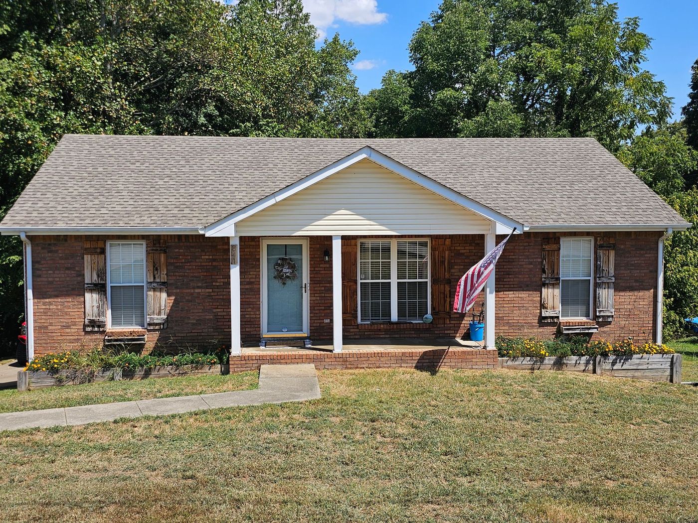 Brick ranch-style house with a blue door and American flag on the porch, surrounded by green grass and trees.