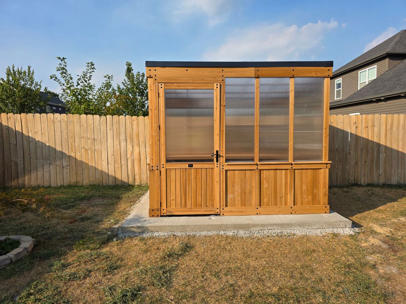 Wooden greenhouse with a door and clear panels, set on a concrete base, beside a wooden fence.