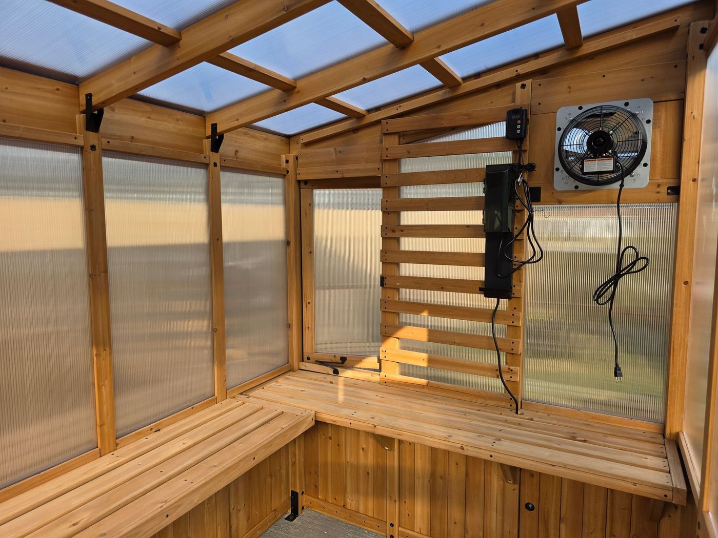Wooden greenhouse interior with benches, fan, and vent panels; lit by sunlight.