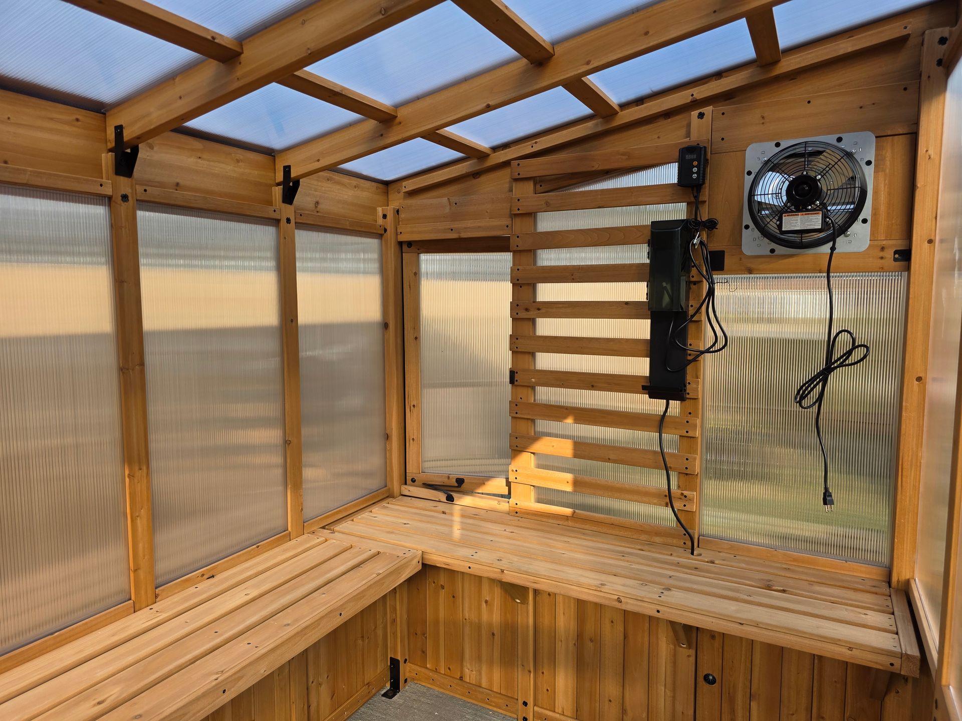 Wooden greenhouse interior with benches, corrugated plastic walls, and a fan.