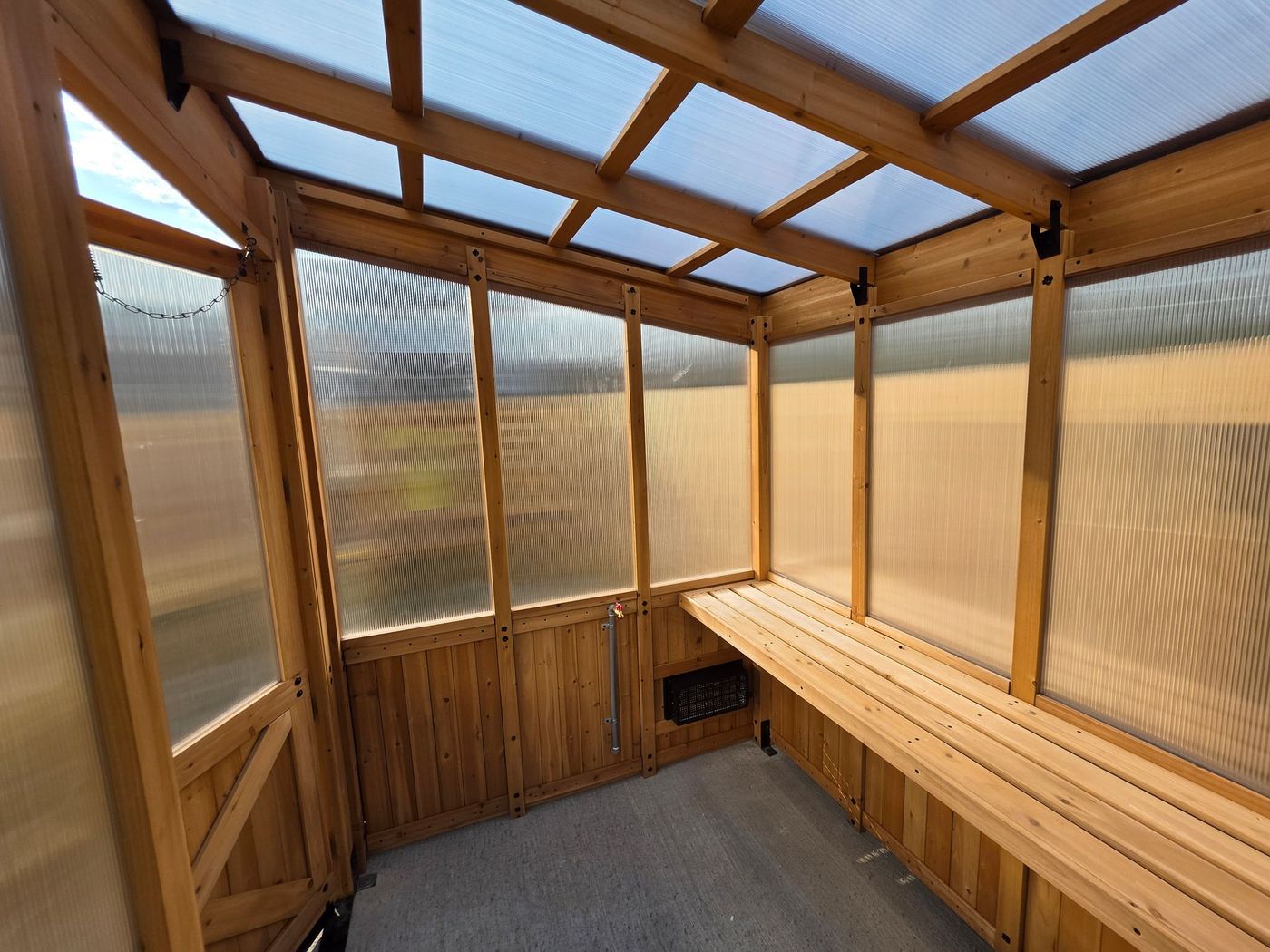 Interior view of a wooden greenhouse with clear plastic panels. Empty benches line the walls under a translucent roof.