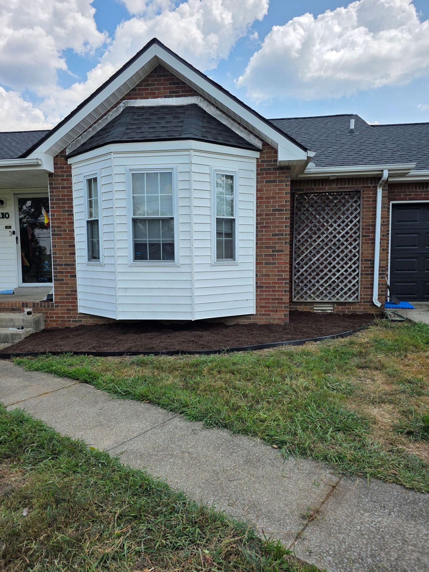 A brick townhome with white bay window and black roof, brown mulch, and a gray walkway.