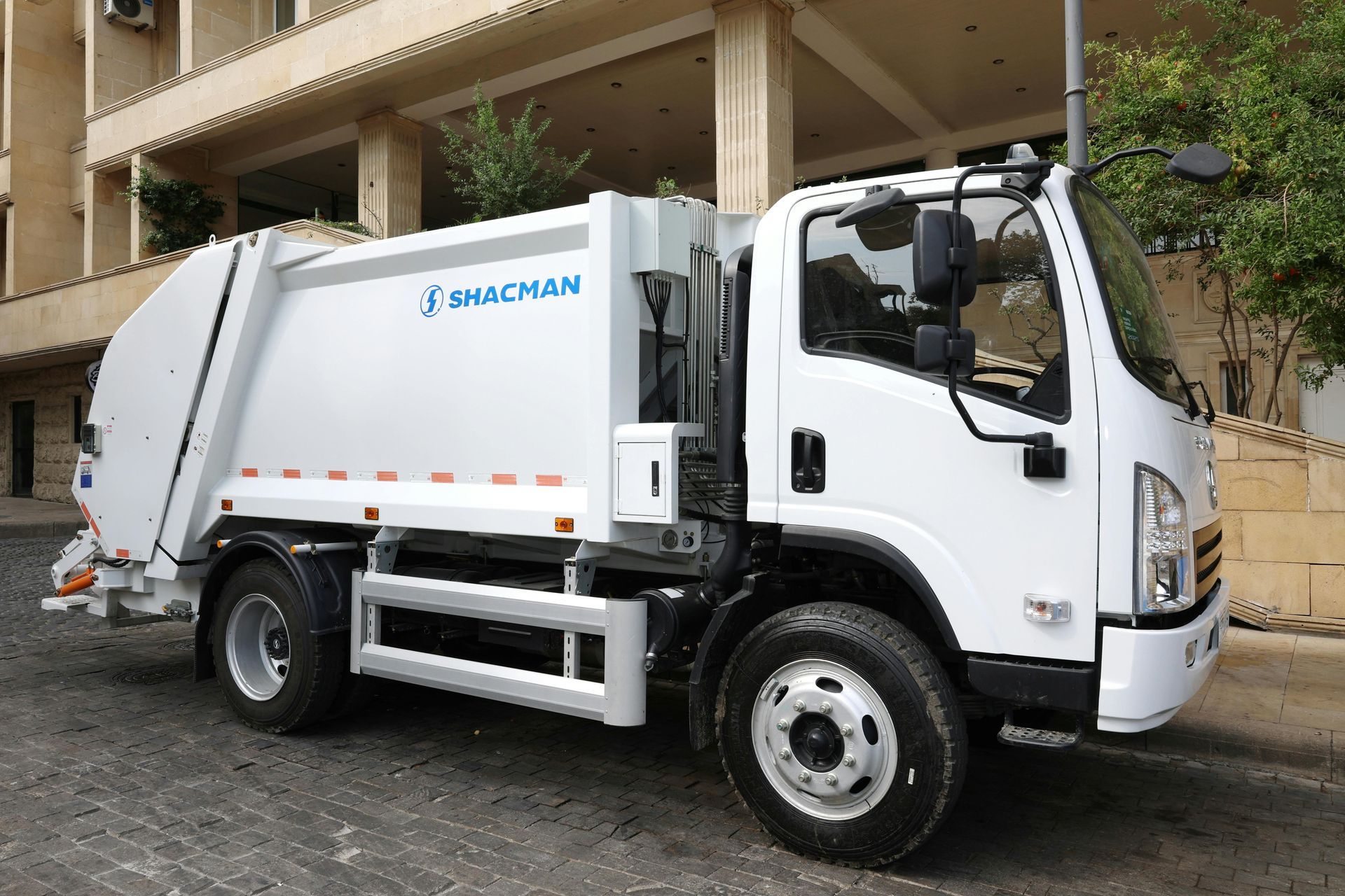 White garbage truck parked on a cobblestone street, in front of a building.