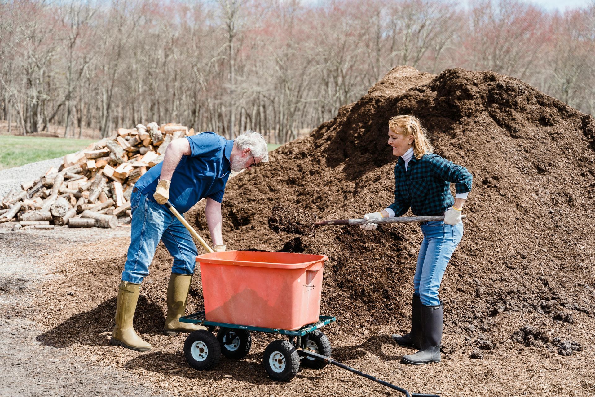 A man and woman load mulch into a wagon near a large pile and woodpile in an outdoor setting.