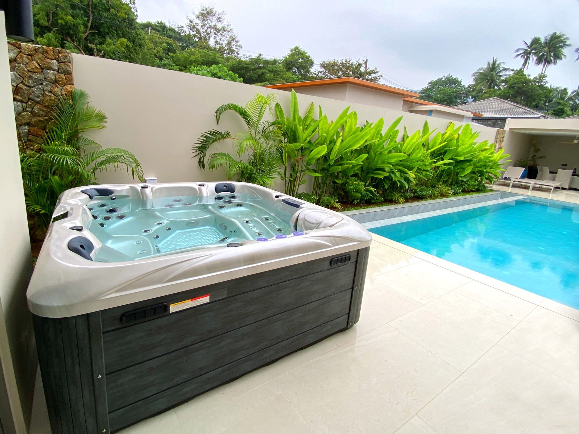 OCEANFRONT Alfa seires - Couple's Hot Tub อ่างน้ำร้อน รุ่น มารีน่า ติดตั้งกลางแจ้งริมสระว่ายน้ำของบ้านพักตากอากาศสไตล์โมเดิร์นในประเทศไทย รายล้อมด้วยสวนเขตร้อน เหมาะสำหรับการพักผ่อนของครอบครัว แช่น้ำอุ่นพร้อมระบบนวด และสลับลงสระน้ำเย็นเพื่อความผ่อนคลาย.