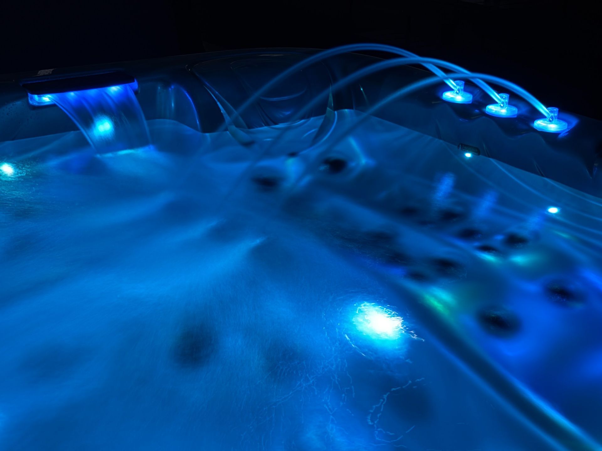 Outdoor hot tub with multi-color LED lighting, waterfall feature, and fountain jets creating a relaxing nighttime ambiance