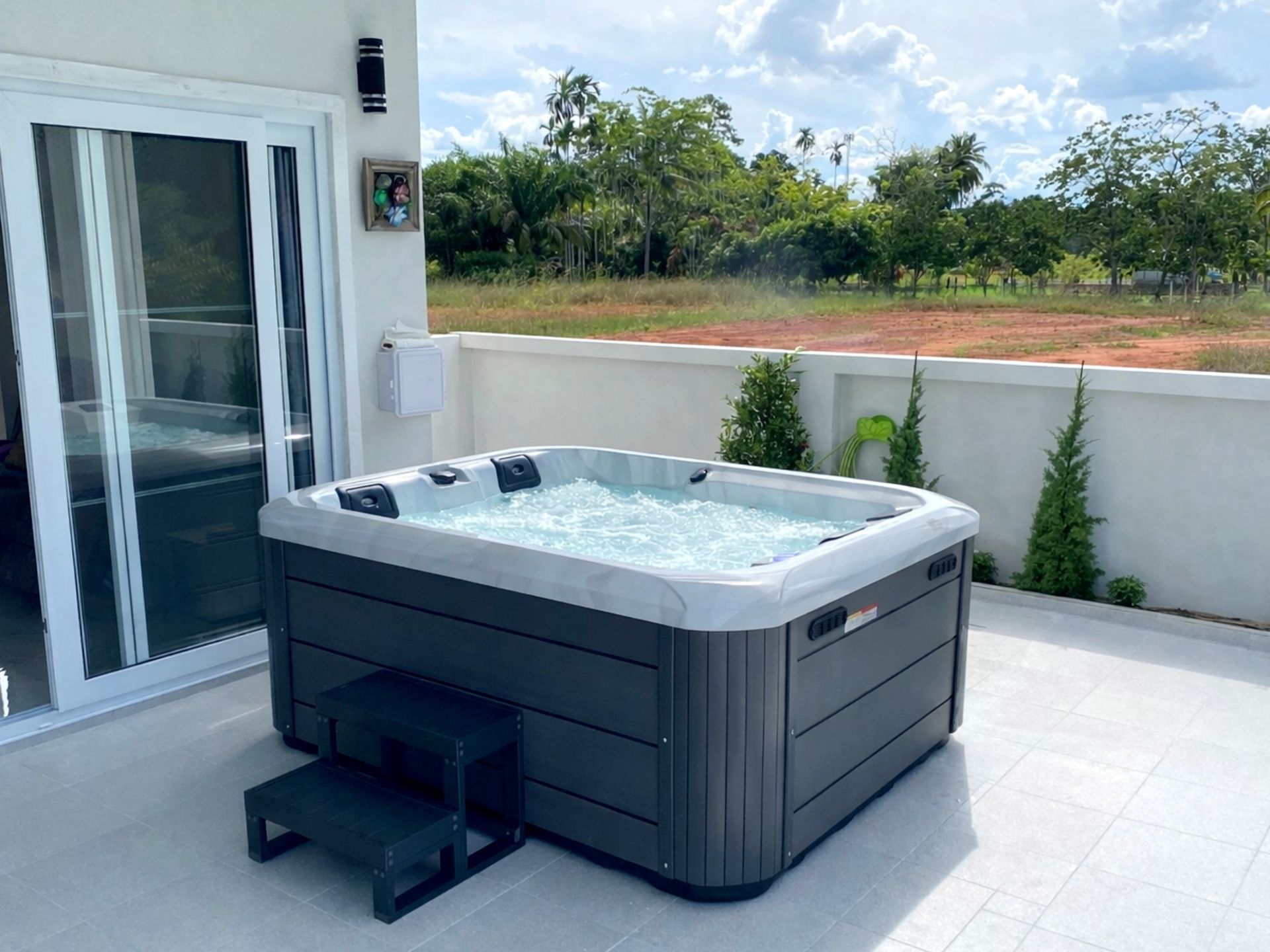 อ่างน้ำร้อนสปากลางแจ้งสีขาวเทา พร้อมระบบเจ็ทนวดน้ำ ติดตั้งบนระเบียงบ้านสไตล์โมเดิร์น เหมาะสำหรับติดตั้งภายนอกบ้านพักอาศัย พร้อมวิวสวนธรรมชาติ เป็นตัวเลือกอ่างน้ำร้อนสำหรับวิลล่า