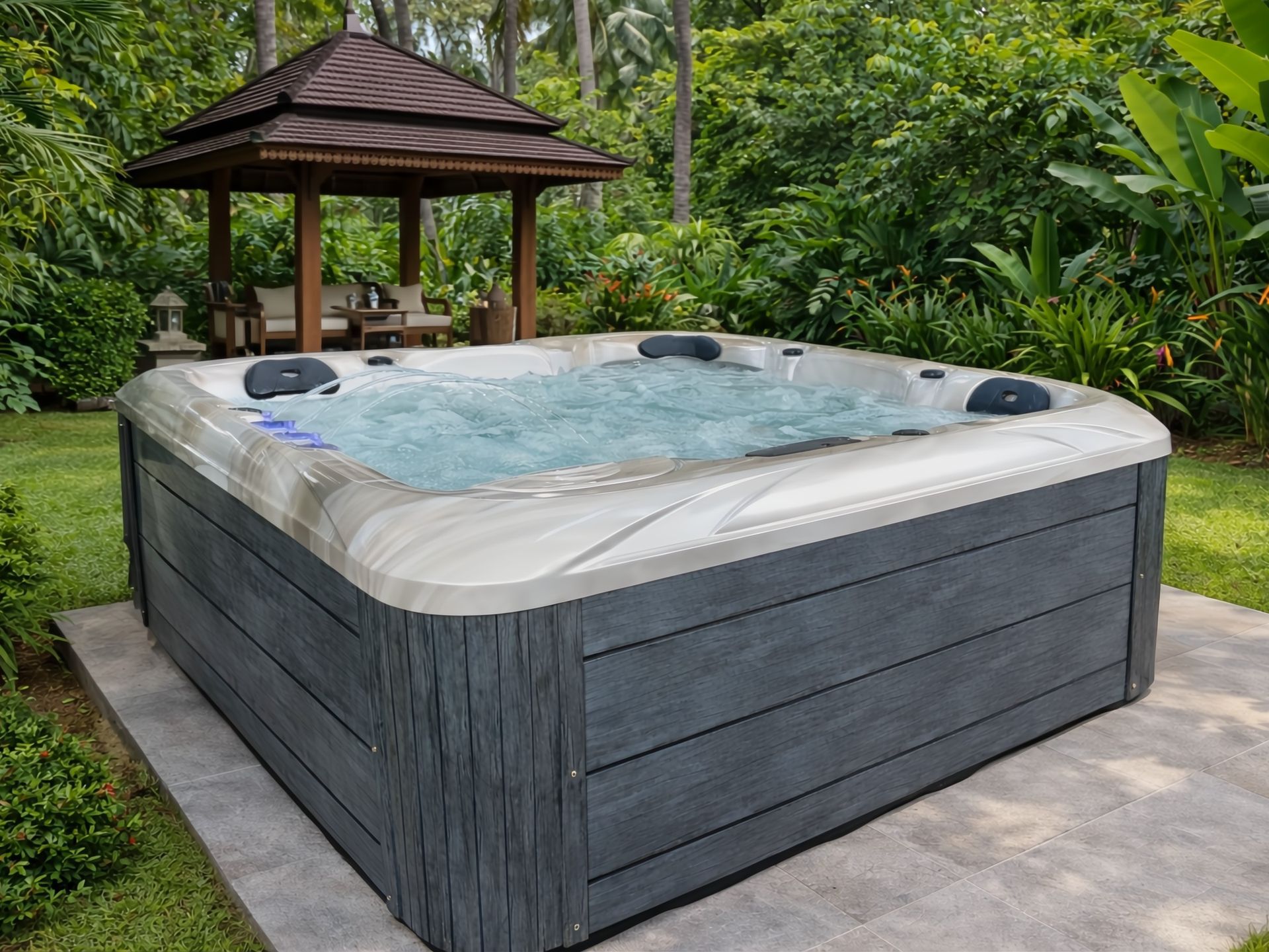 MARINA 5-person outdoor hot tub with 2 loungers and 3 seats installed in a tropical garden setting near a wooden pavilion, creating a private resort-style spa atmosphere.