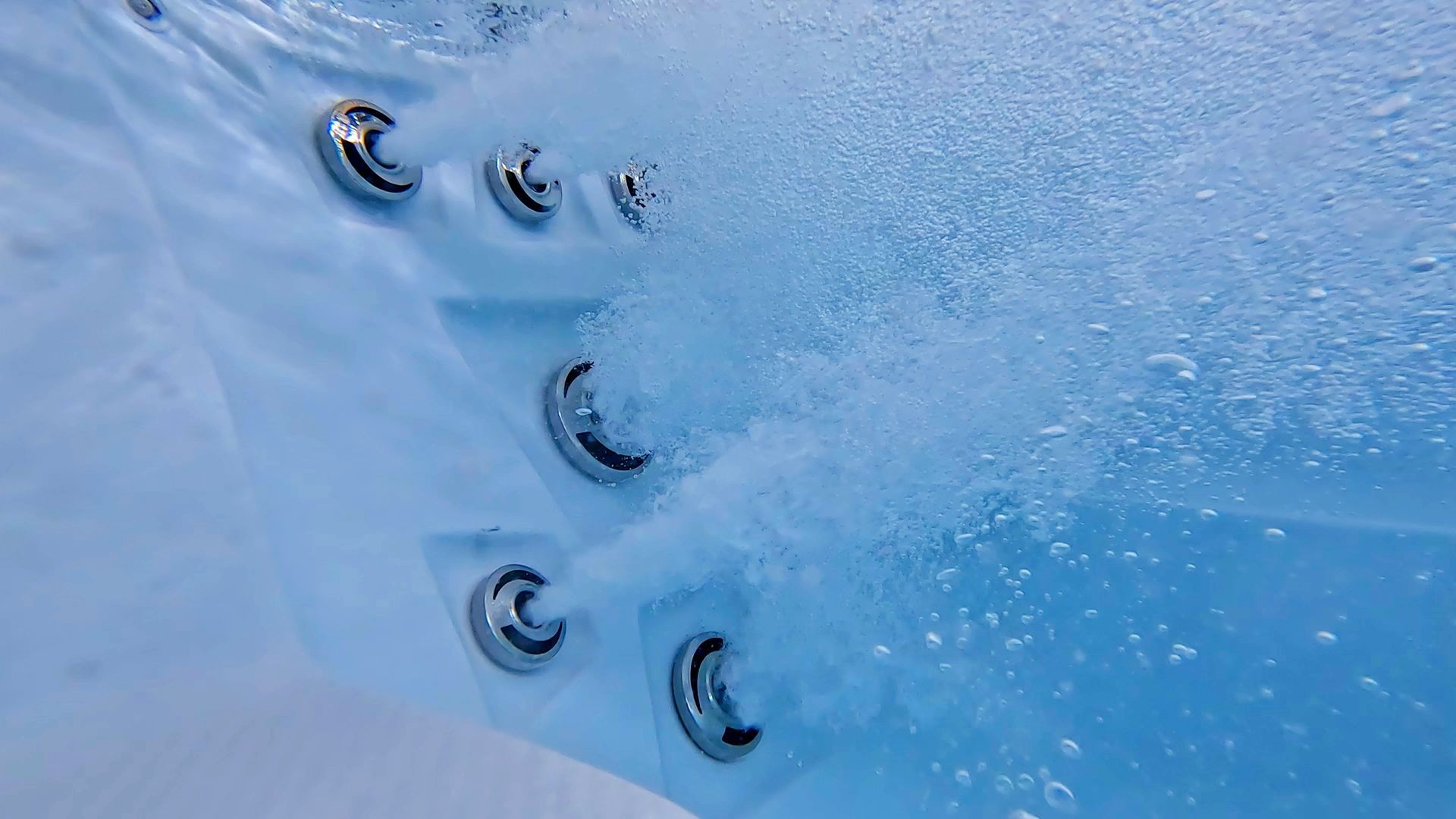 Underwater view of a hot tub wall with multiple metal jets shooting streams of bubbles into clear blue water